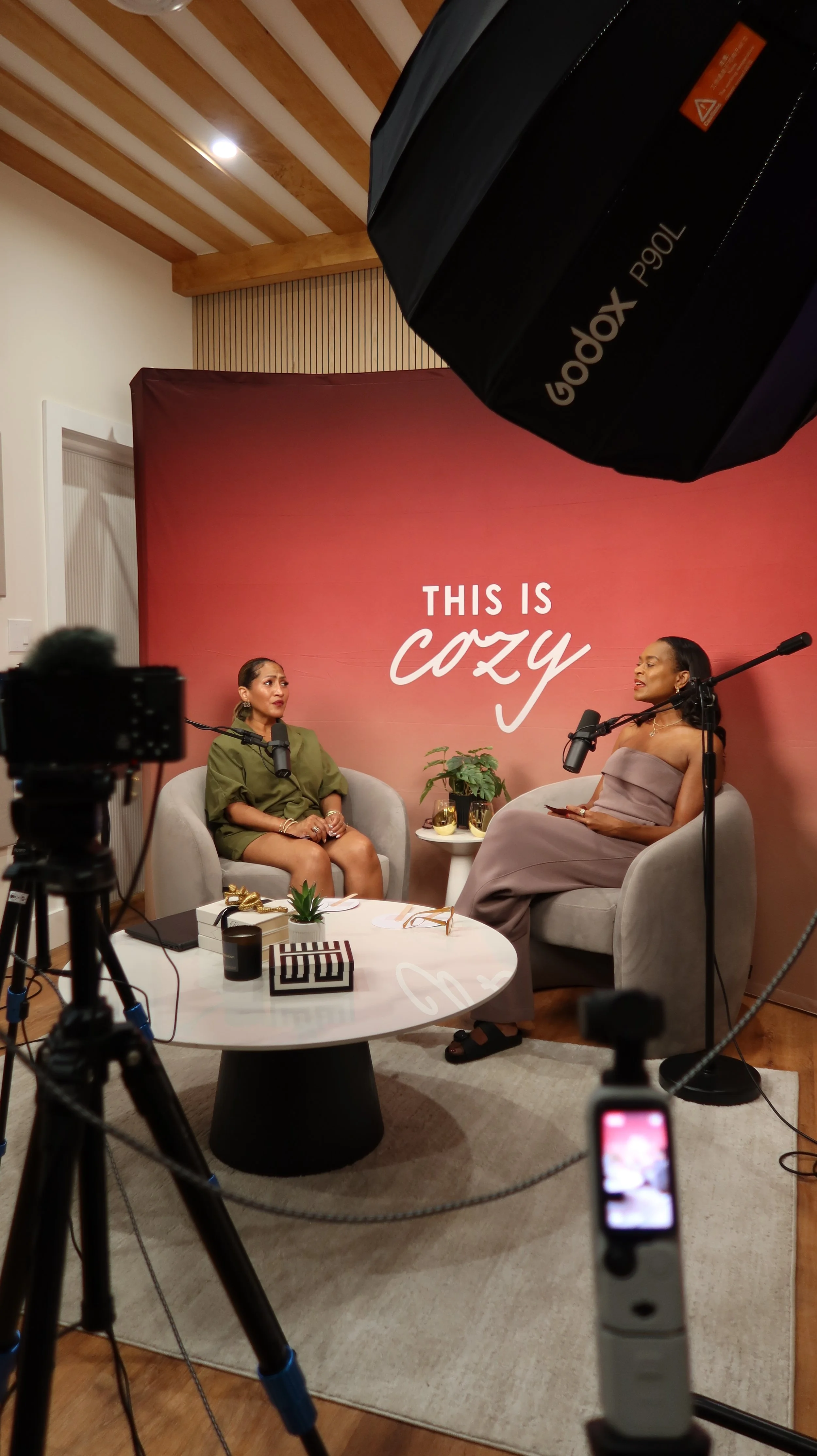 Two women are sitting on gray armchairs in a cozy interview setting, with a red backdrop displaying the words "This is Cozy." They are surrounded by professional camera equipment, including a large softbox and a tripod. A small white table between them holds decorative items like a potted plant, wine glasses, and a box.