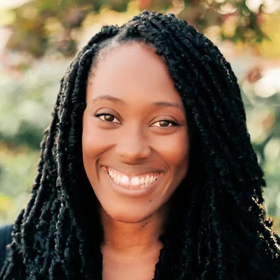 A smiling woman with dark skin and black twisted hair outdoors with a blurred natural background.