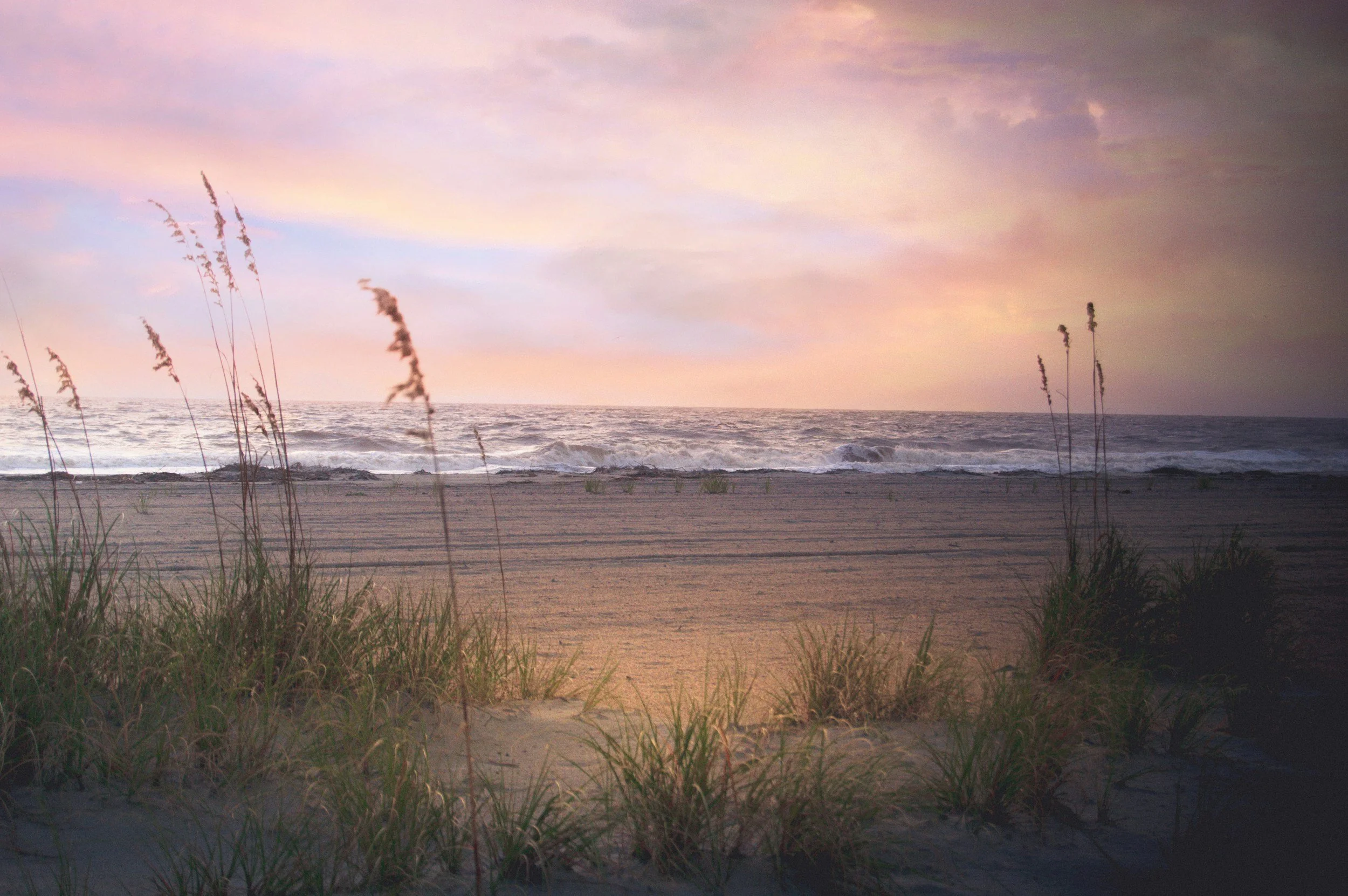 A beach scene at sunset with tall grass in the foreground, waves crashing on the shore, and a colorful sky with pink and purple hues.