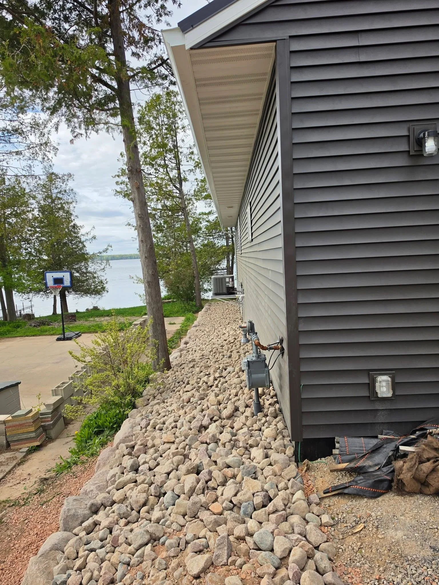 Side of house gravel around basketball hoop — River rock and gravel installation along house exterior by Ground & Grid Solutions 