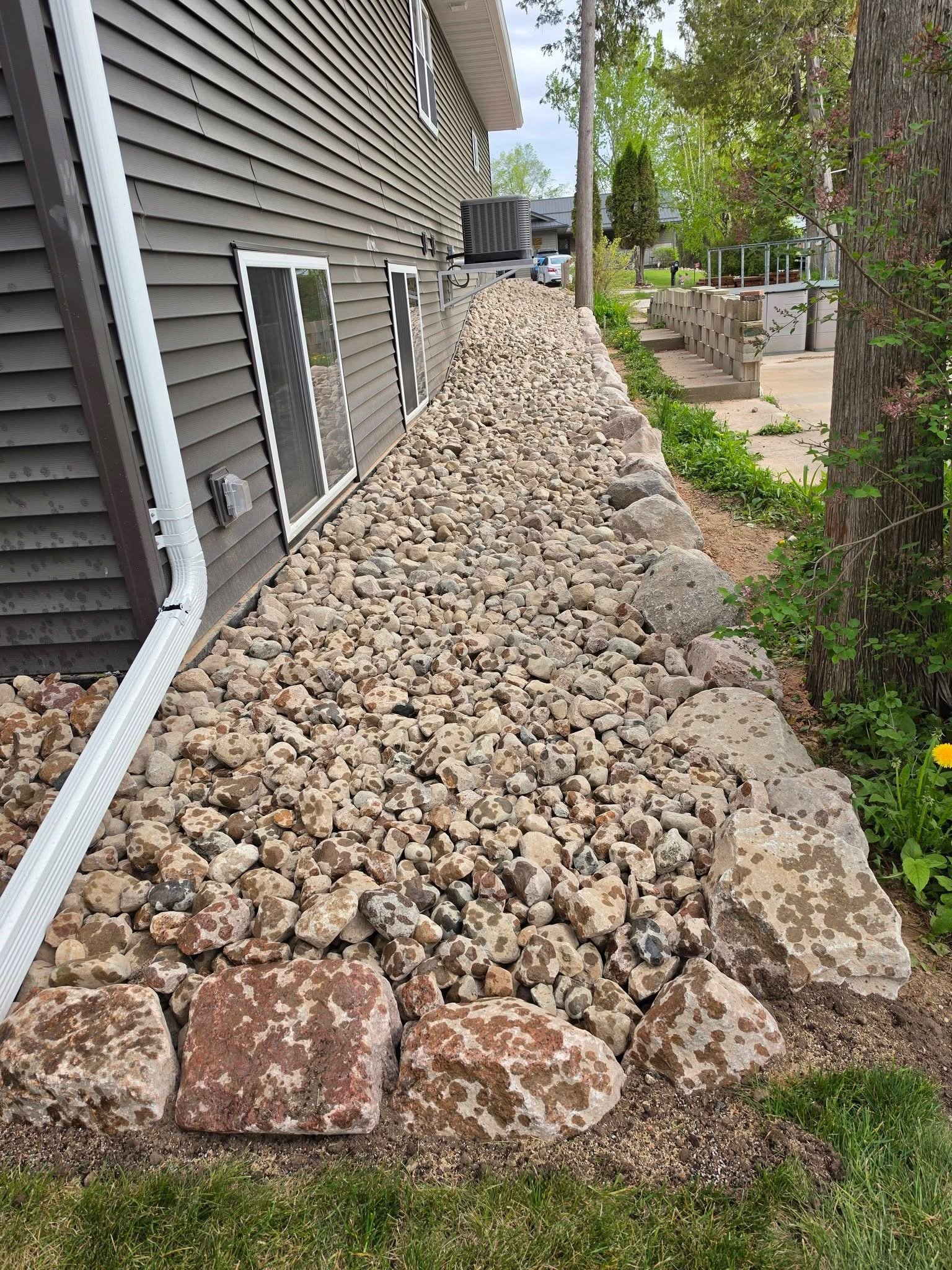 Large rocks house base — River rock drainage bed and stone placement along house foundation by Ground & Grid Solutions