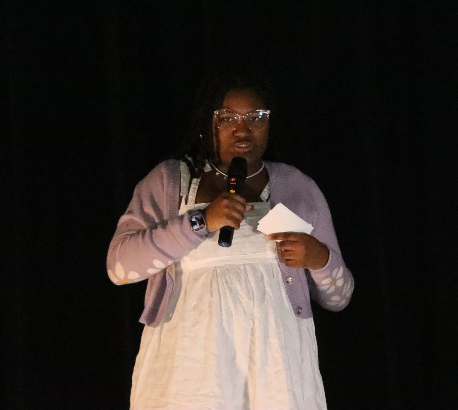 A young woman holding a note card speaks into a microphone on a dark stage
