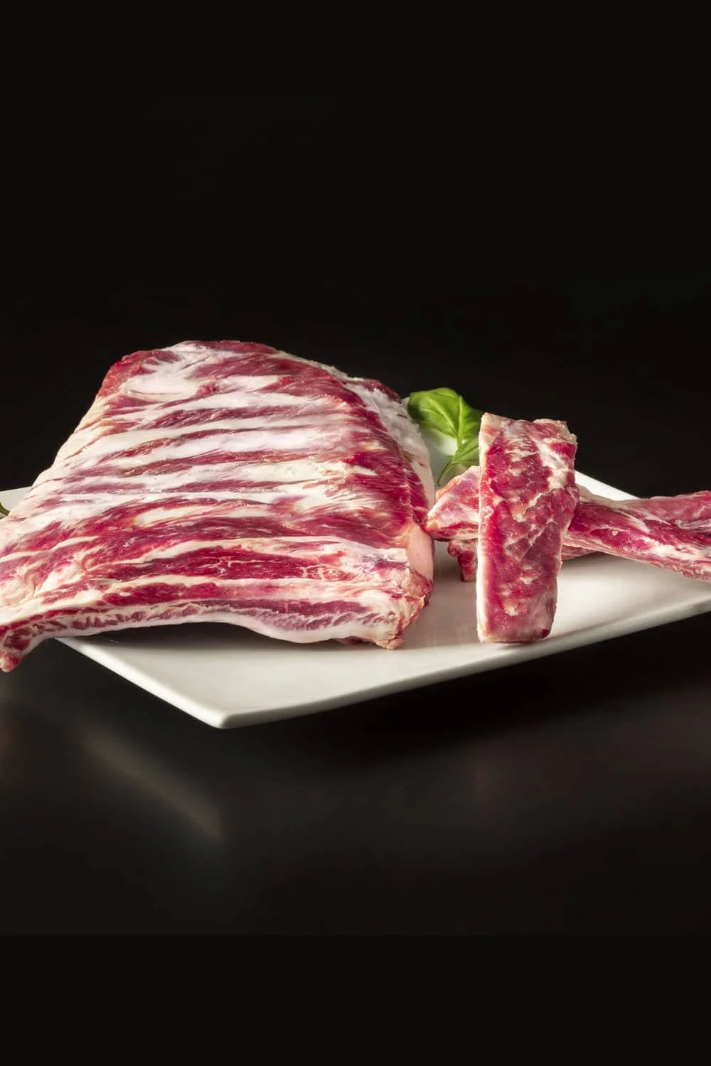 Cesar Nieto Iberico Pork Short Ribs finger (cubes) on a white plate, garnished with a small piece of green leaf, against a black background.