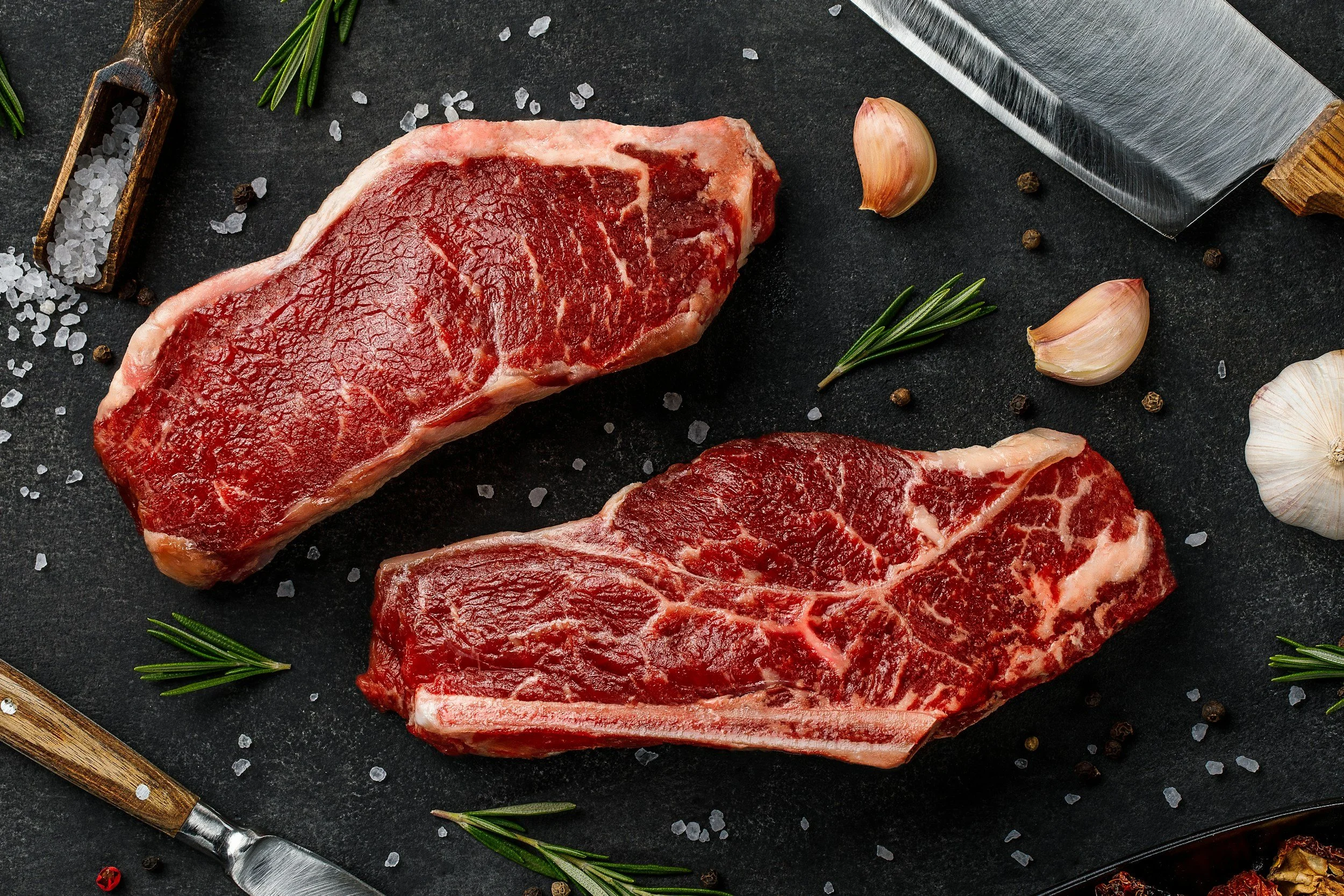 Two raw Striploin beef steaks on a black surface with coarse salt, garlic cloves, black peppercorns, rosemary sprigs, and a large knife.