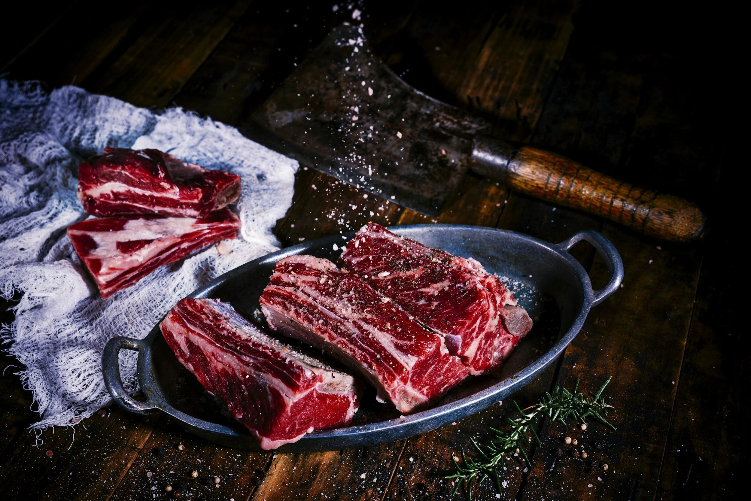 Raw Short Rib beef steaks on a rustic wooden table with salt being sprinkled on them, surrounded by herbs and seasonings.