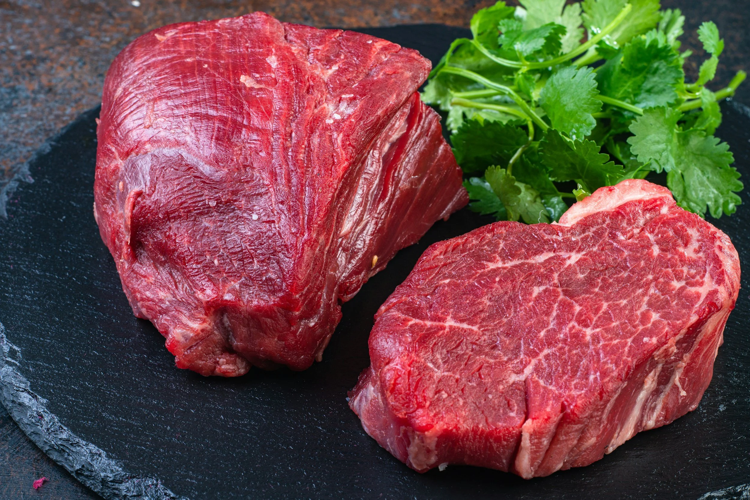 Two pieces of raw Zabuton (Chuck Flap Tail) beef, on a black slate plate accompanied by green parsley garnish.
