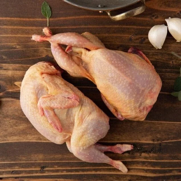 Two raw whole French Quail on a wooden surface, with garlic cloves and a bay leaf nearby.