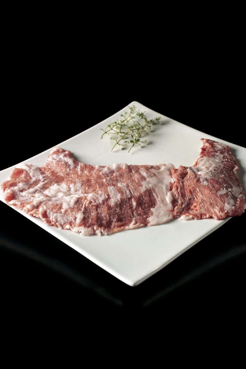 Cesar Nieto Iberico Pork Jowl Secreto on a white square plate with a sprig of thyme, set against a black background.