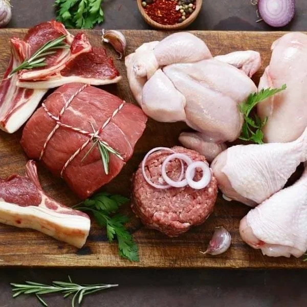 Assorted raw meats including beef, chicken, and ground meat with onion slices, garlic, herbs, and spices on a wooden cutting board.