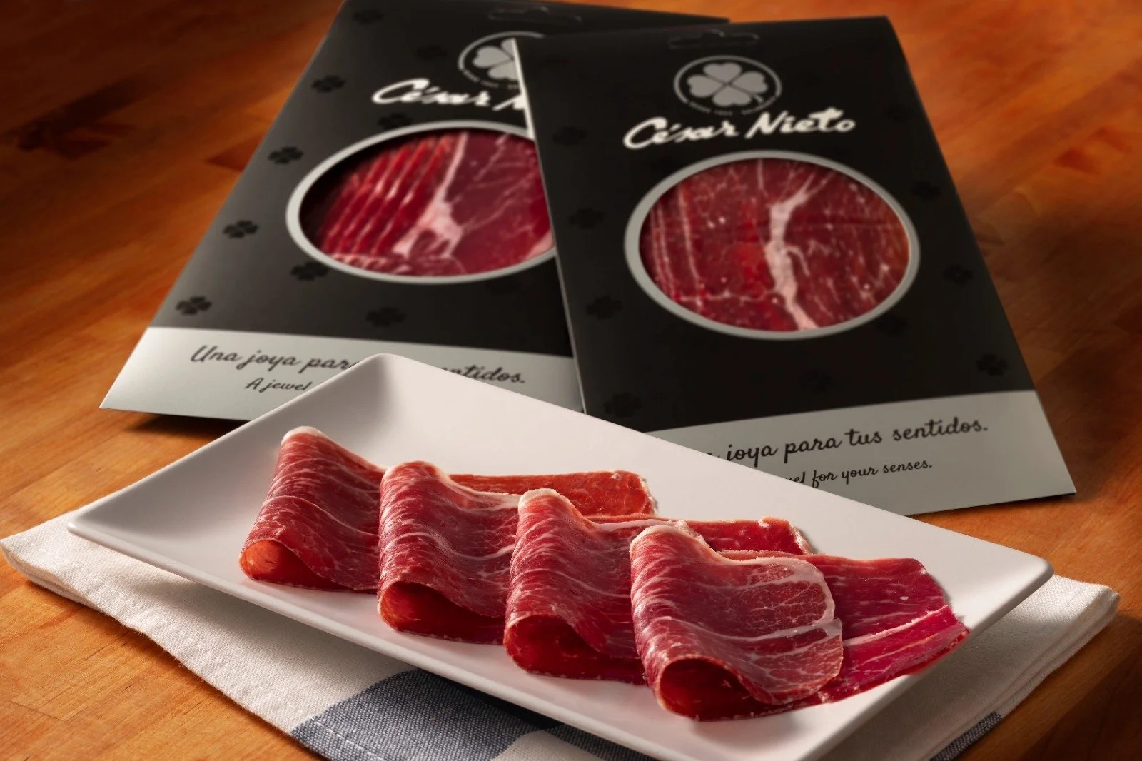 Slices of Spanish Spanish "César Nieto" Iberico ham on a white rectangular plate, placed on a cloth napkin on a wooden table. Behind the plate are two packages of sliced cured ham with black packaging and circular windows showing the meat inside.