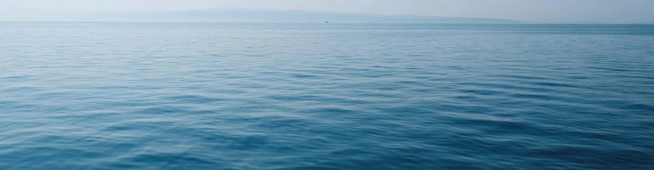 Calm ocean water with gentle waves, a hazy horizon, and distant land or mountain silhouette.