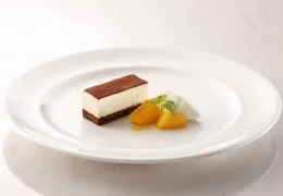 A slice of Japan Saveur Tiramisu Short Cake with a dark top layer, creamy middle layer, and light base, served with sliced peaches and a sprig of mint on a white plate.