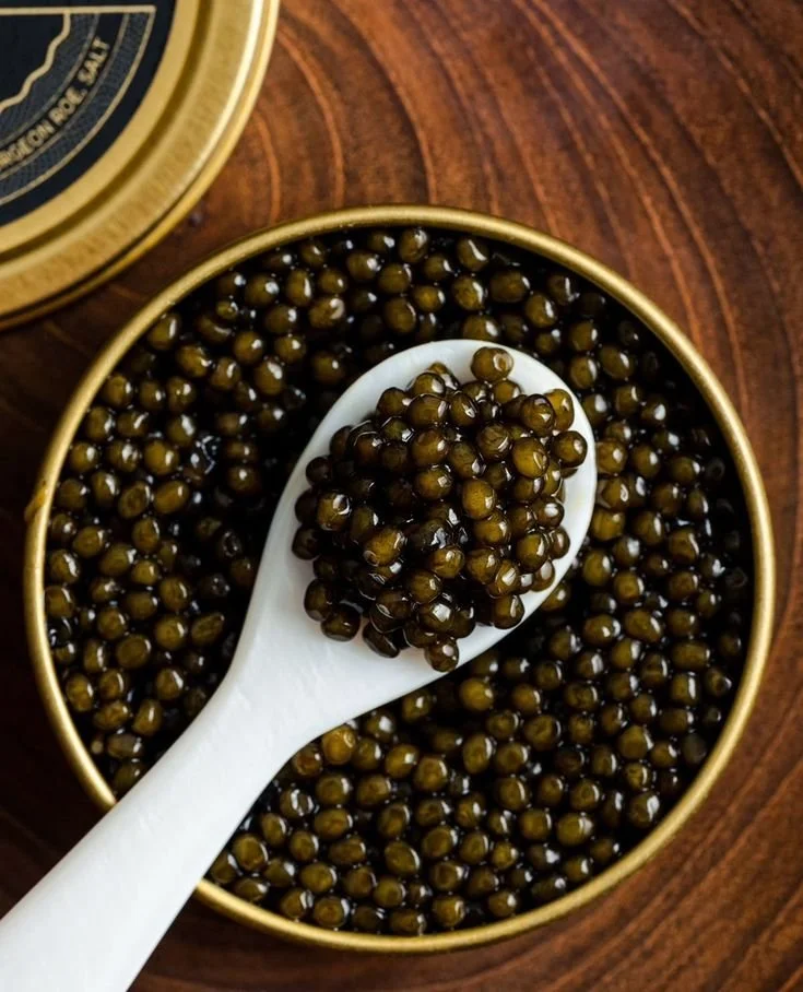 Close-up of a white spoon filled with Oscetra Caviar, placed inside a round golden tin filled with more black caviar, on a wooden surface.