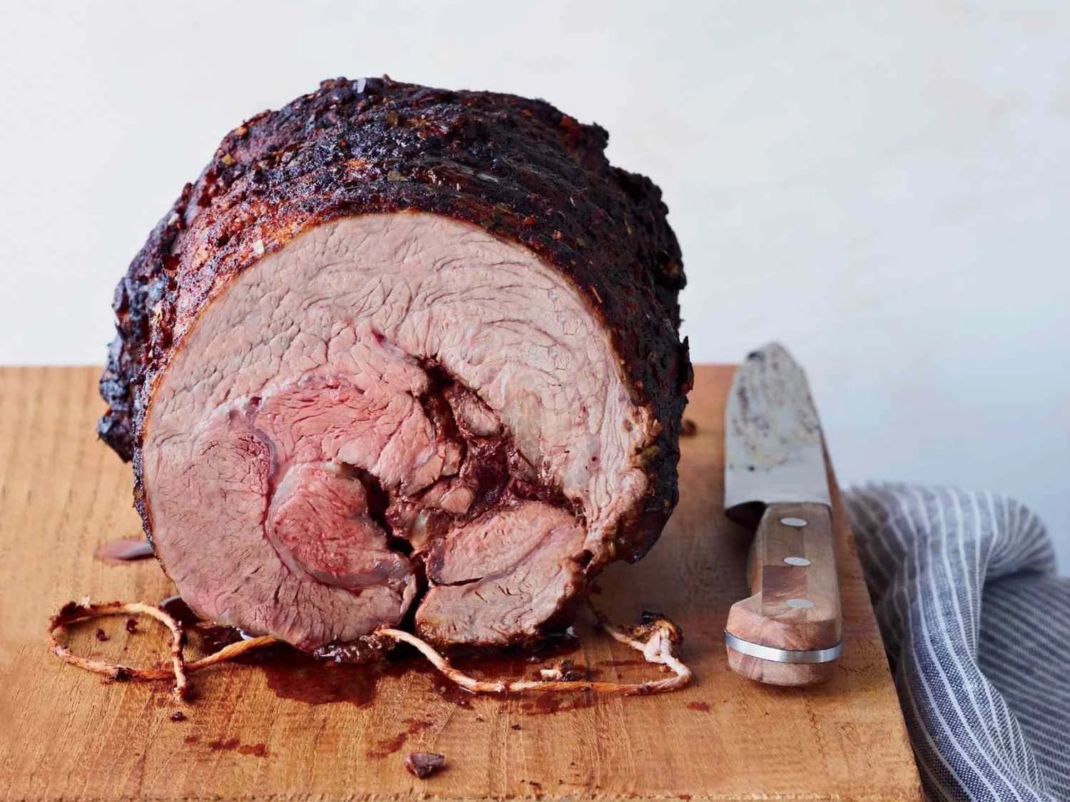 A large rolled Chuck Eye Roll beef roast with a dark, crispy outer crust on a wooden cutting board, with a carving knife and a striped kitchen towel beside it.