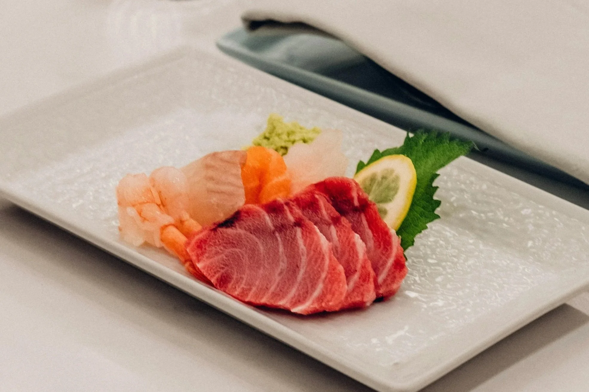 A white rectangular plate with slices of Philippines Yellowfin Tuna Saku, garnished with a lemon wedge, wasabi, and a green leaf. A black utensil is resting above the plate.