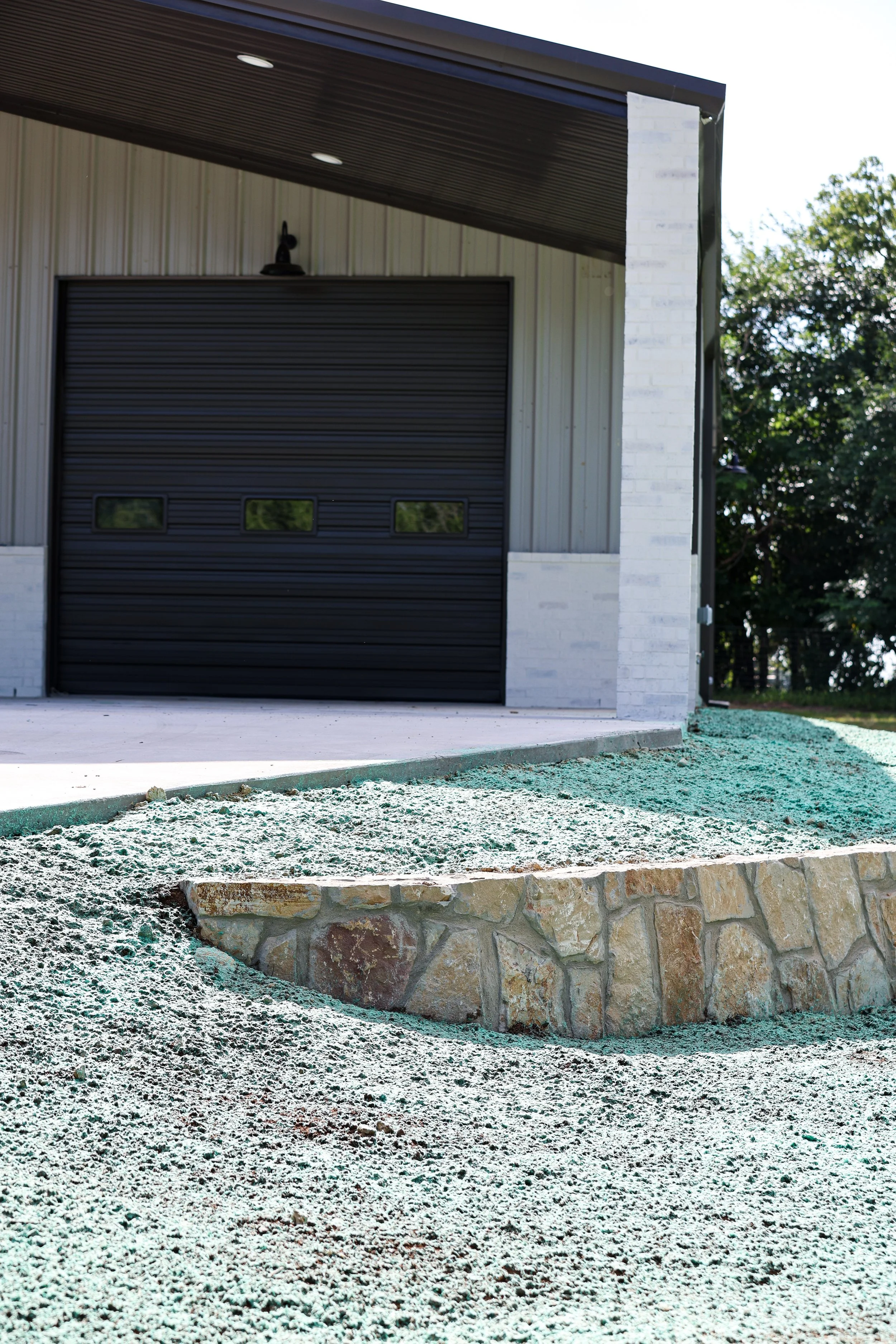 Exterior of a building with a black garage door, a white brick pillar, and trees in the background. The ground shows a partially constructed or landscaped area with dirt and stone borders.