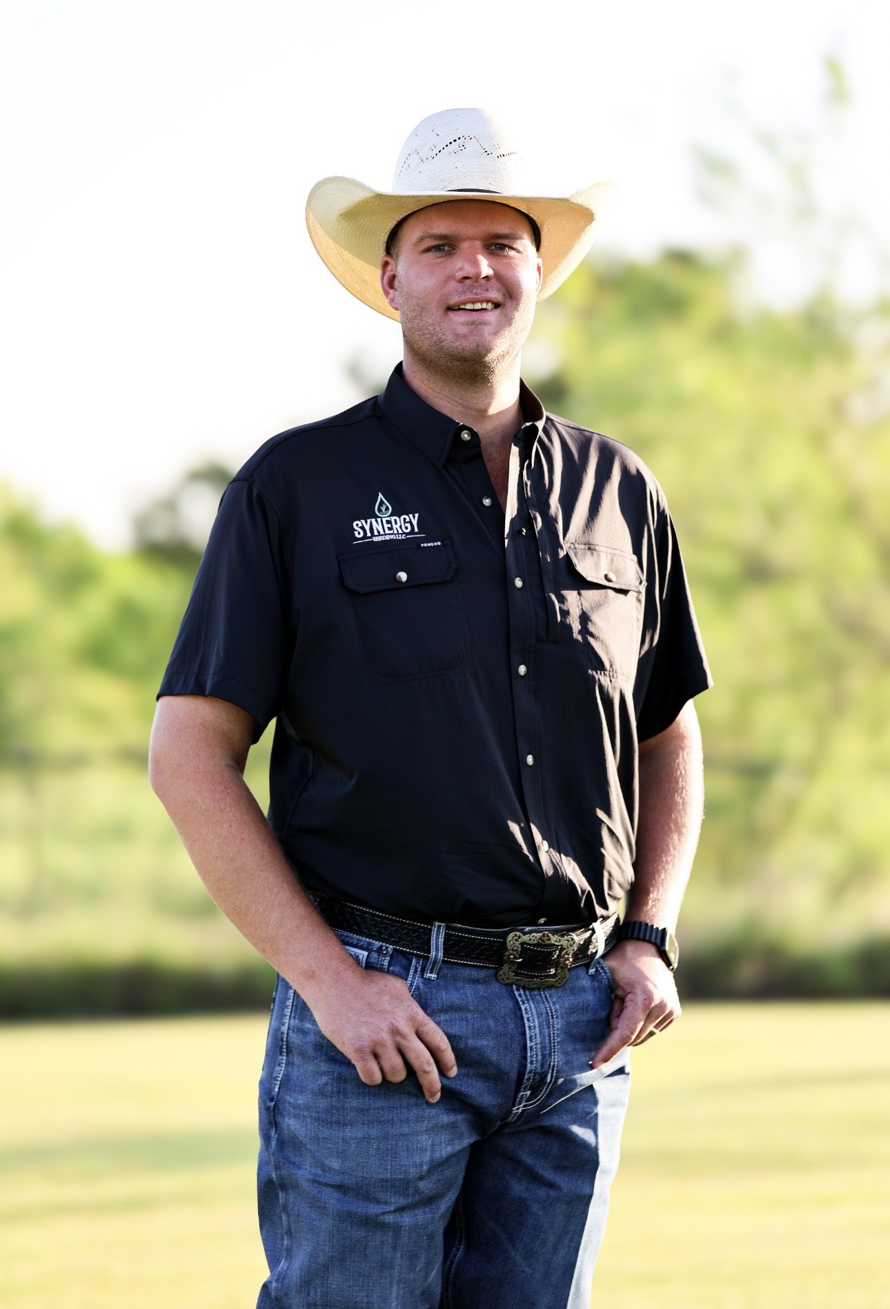 A man wearing a cowboy hat and a black shirt with 'Synergy' logo standing outdoors on a sunny day.