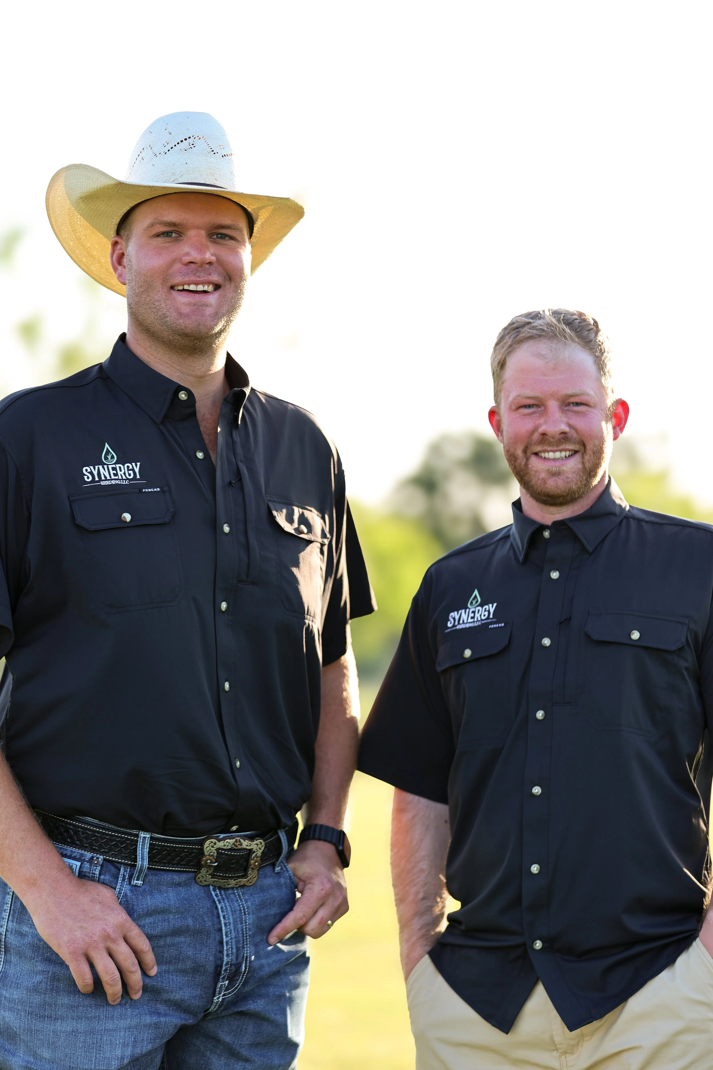 Two men standing outdoors, smiling, wearing black shirts with logos. One man has a cowboy hat, blue jeans, and a belt with a large buckle. The other man has short curly hair and with a beard, wearing khaki pants.