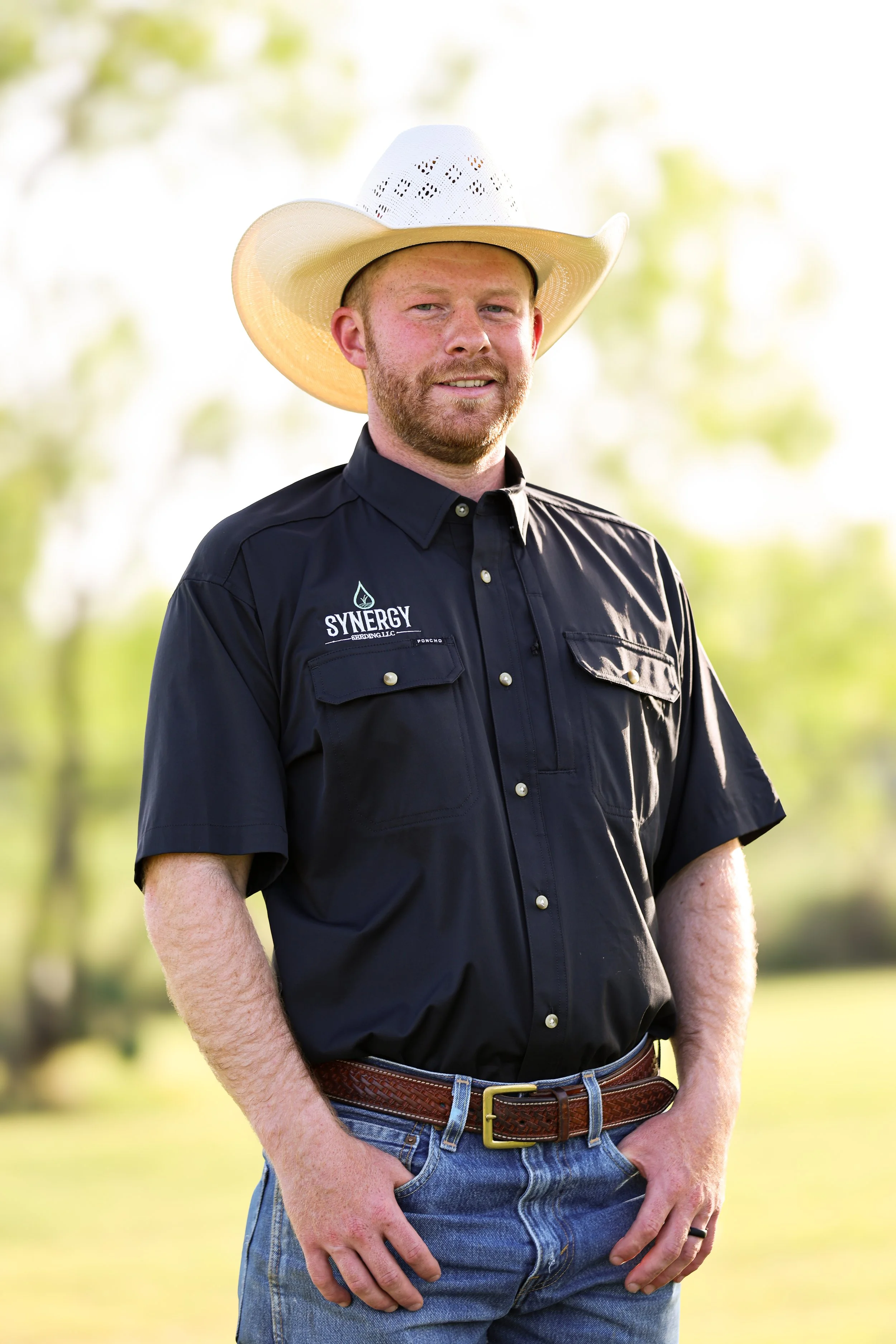 A man wearing a white cowboy hat, black shirt with a logo that says "SYNERGY," blue jeans, and a brown woven belt standing outdoors with a blurred green background.