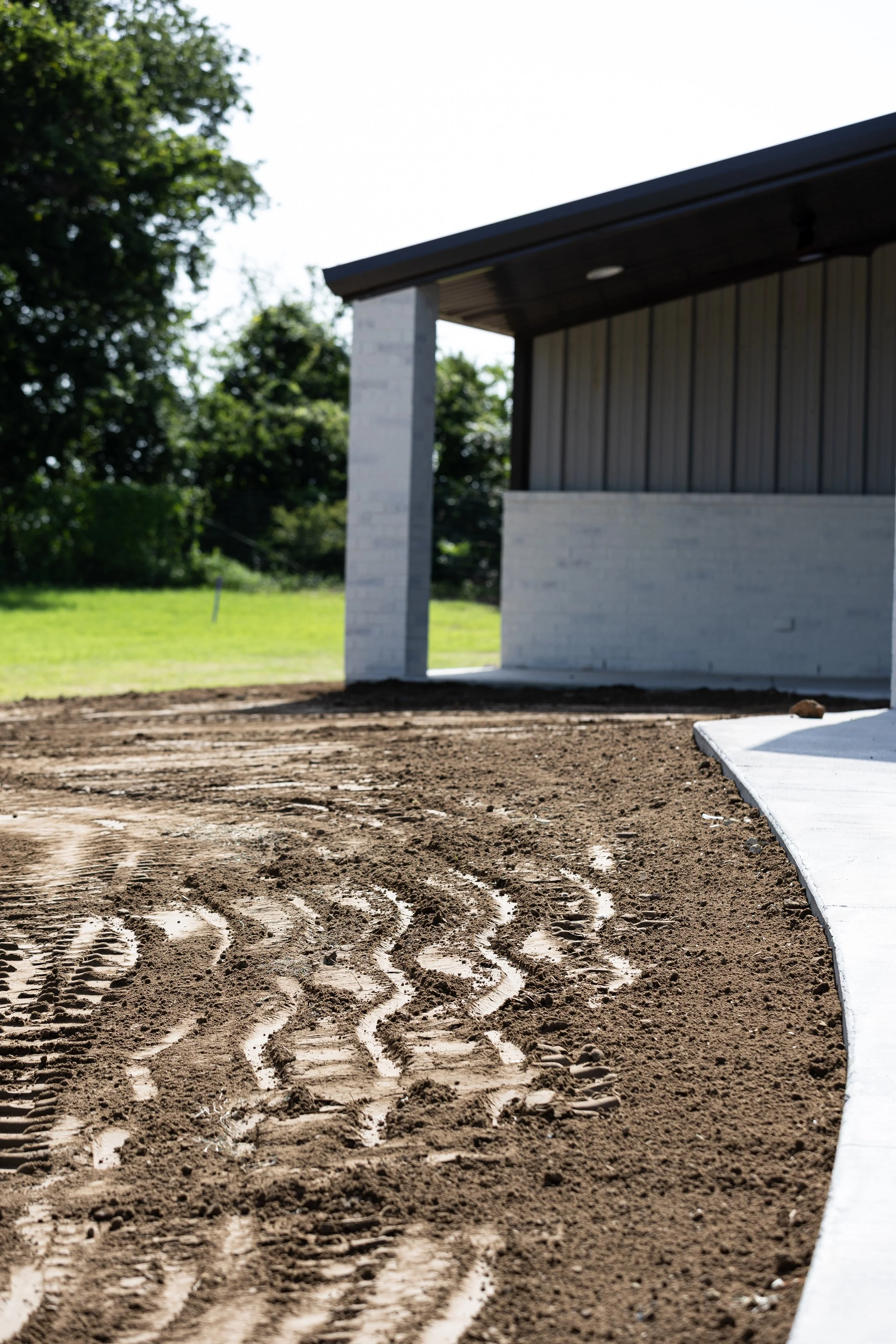 A building under construction with a concrete foundation and dirt ground, tire tracks from construction vehicles, and a small section of a concrete sidewalk. The building has gray siding and a dark roof, and there are trees in the background.