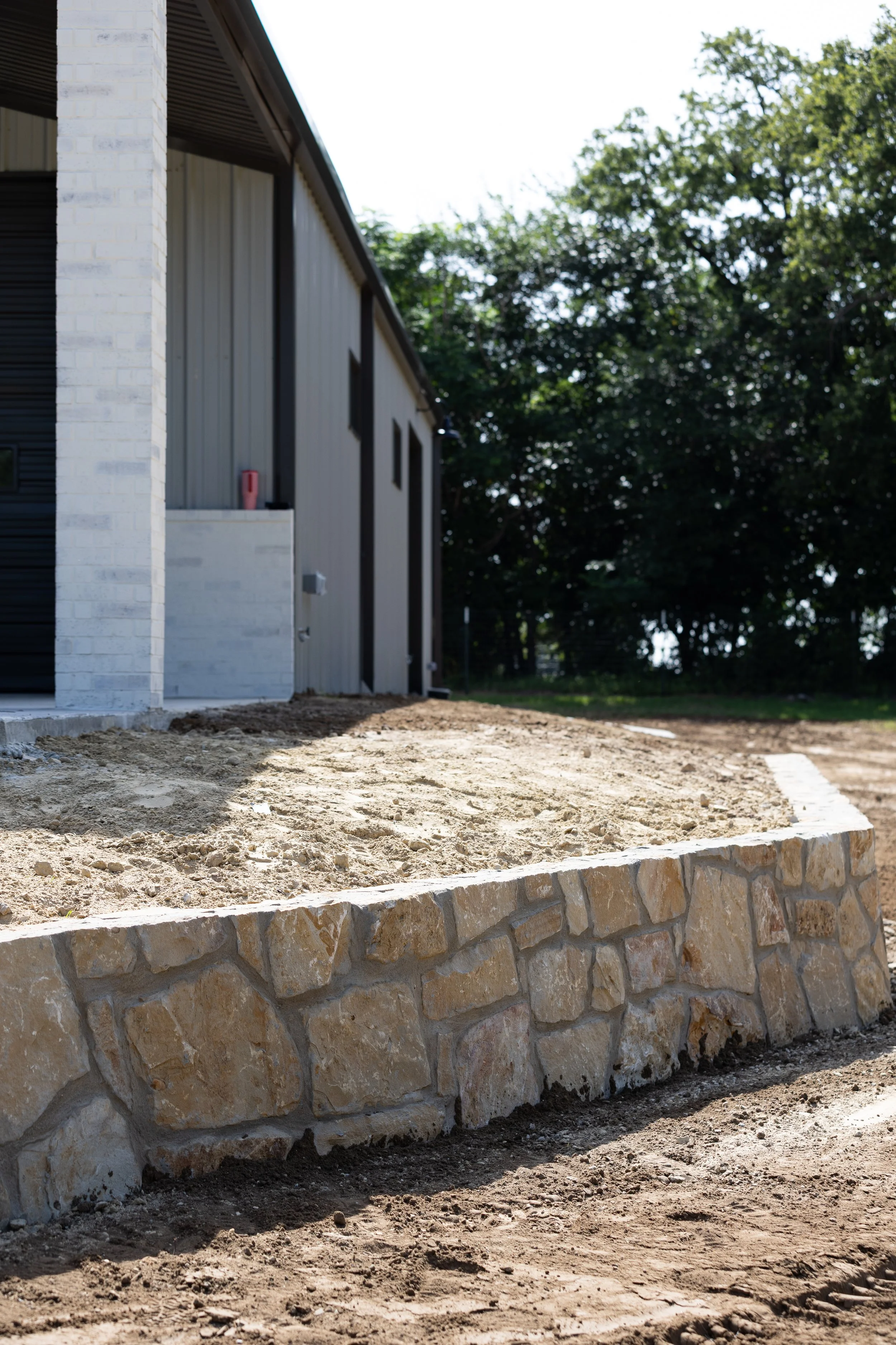 A building under construction with a stone retaining wall in the foreground, dirt ground, and trees in the background.