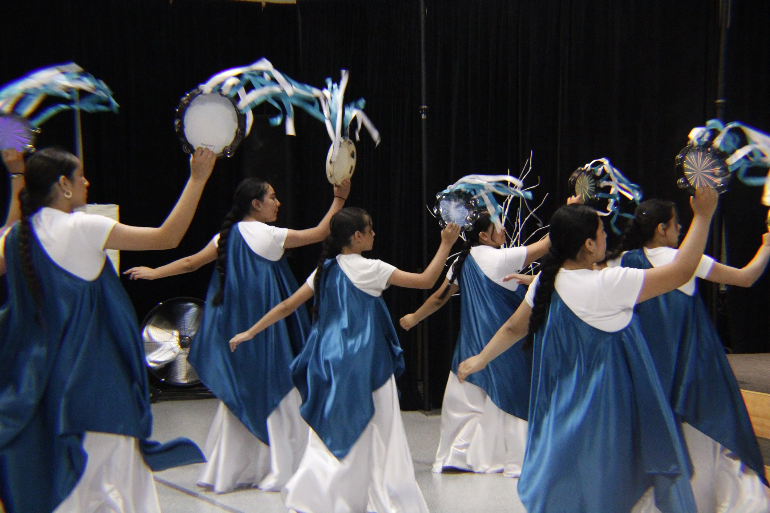A group of people dressed in blue dresses and white shirts, holding tambourines decorated with blue and white ribbons, performing a dance in a line in front of a black curtain.