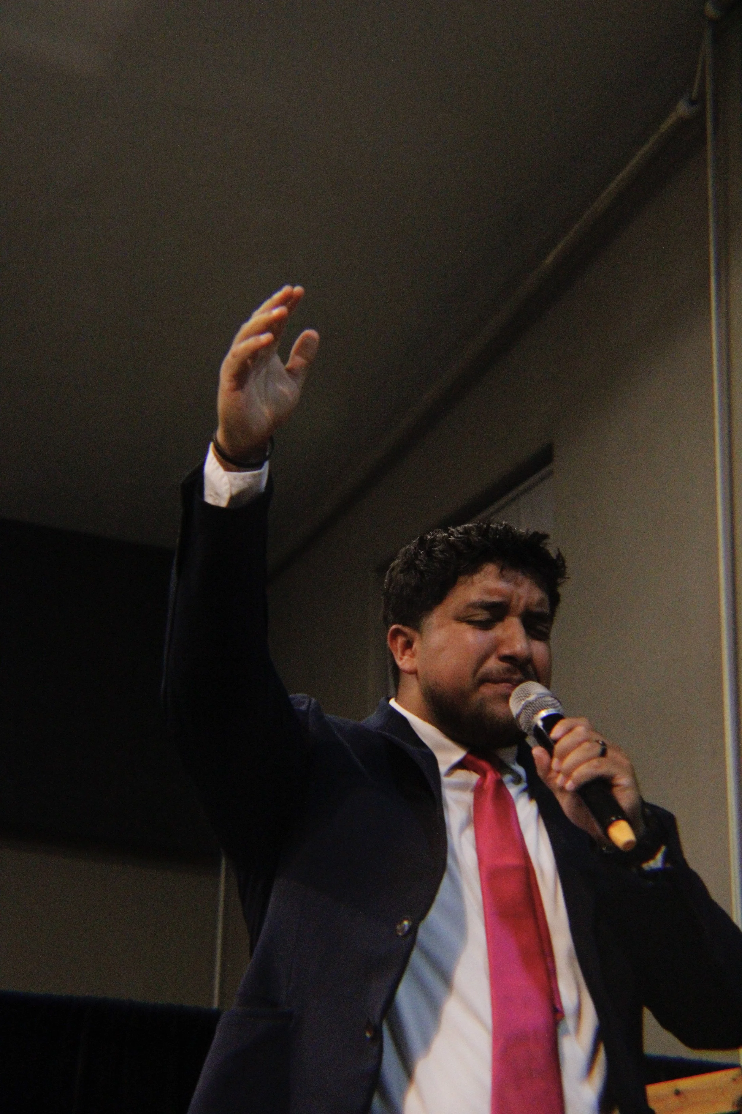 A man in a dark suit and pink tie holding a microphone and raising his hand during a speech or presentation.
