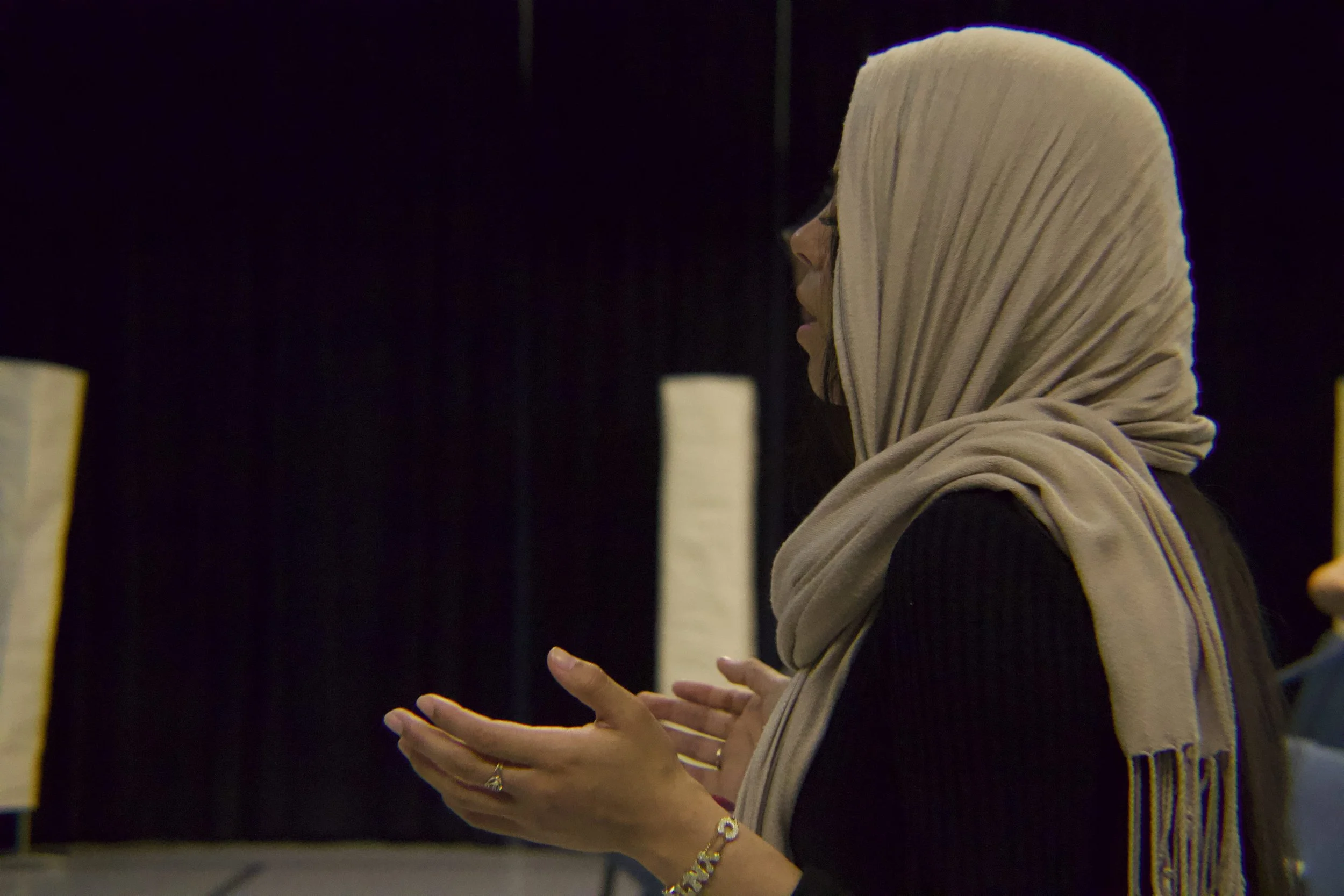 A woman wearing a beige headscarf and glasses, standing against a dark background, appears to be in a contemplative or speaking pose with hands clasped.