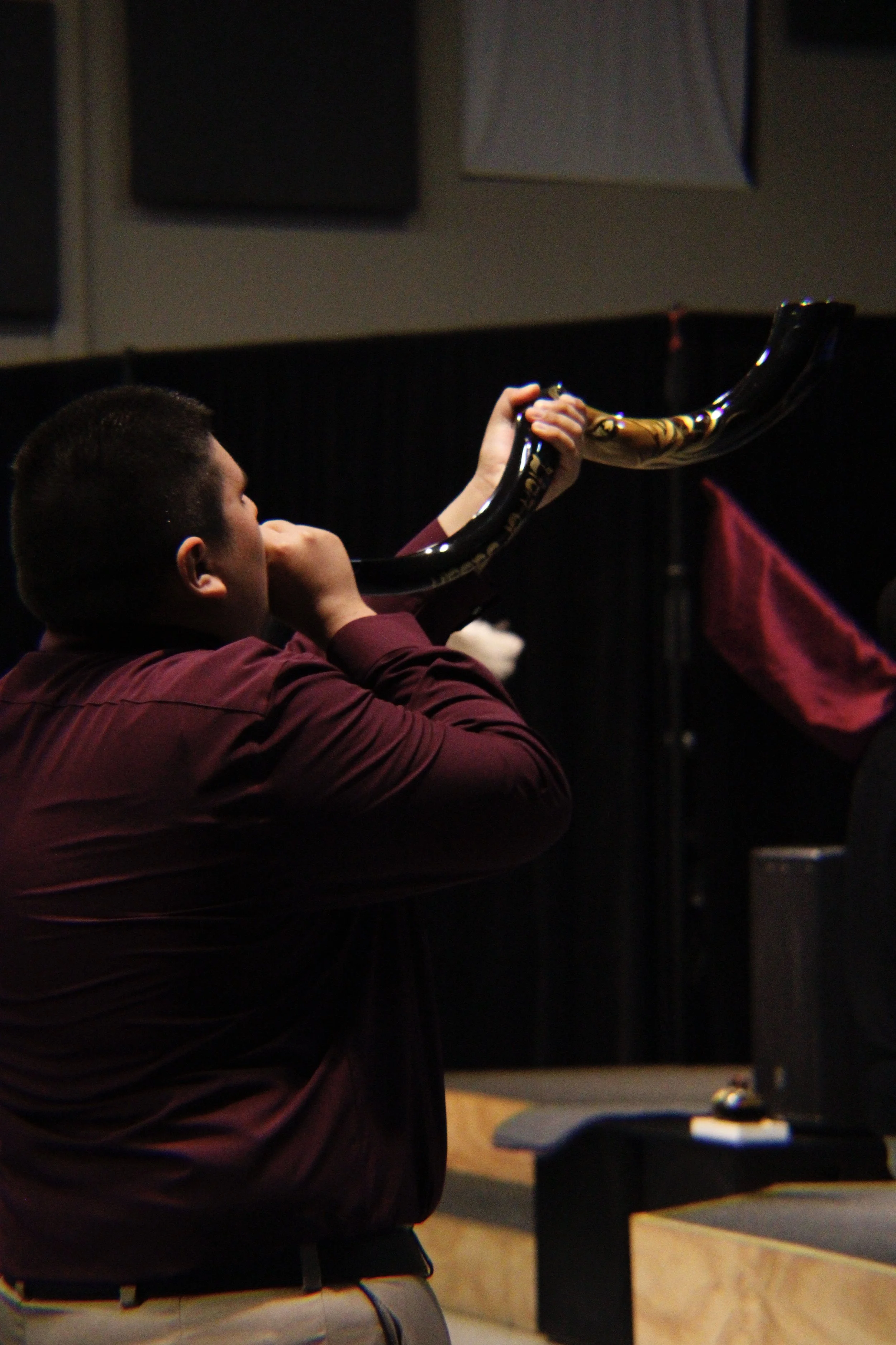 A young man with short hair, wearing a maroon shirt and beige pants, is sitting and playing a large ceremonial horn in a dark room, possibly a church or ceremonial hall.