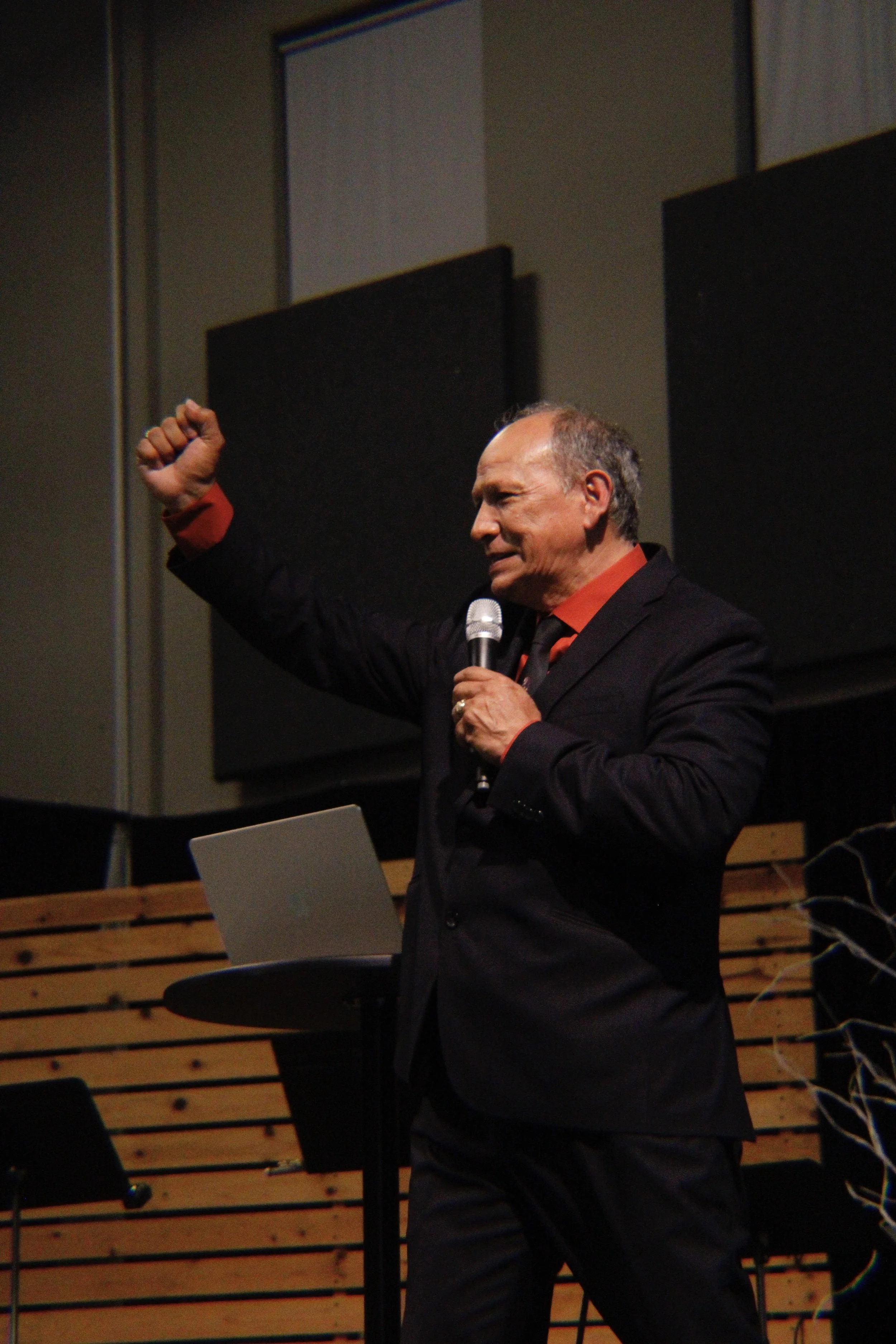 An older man in a black suit with red shirt and tie, holding a microphone, raising his right fist during a presentation or speech, with a laptop and music stands in the background.