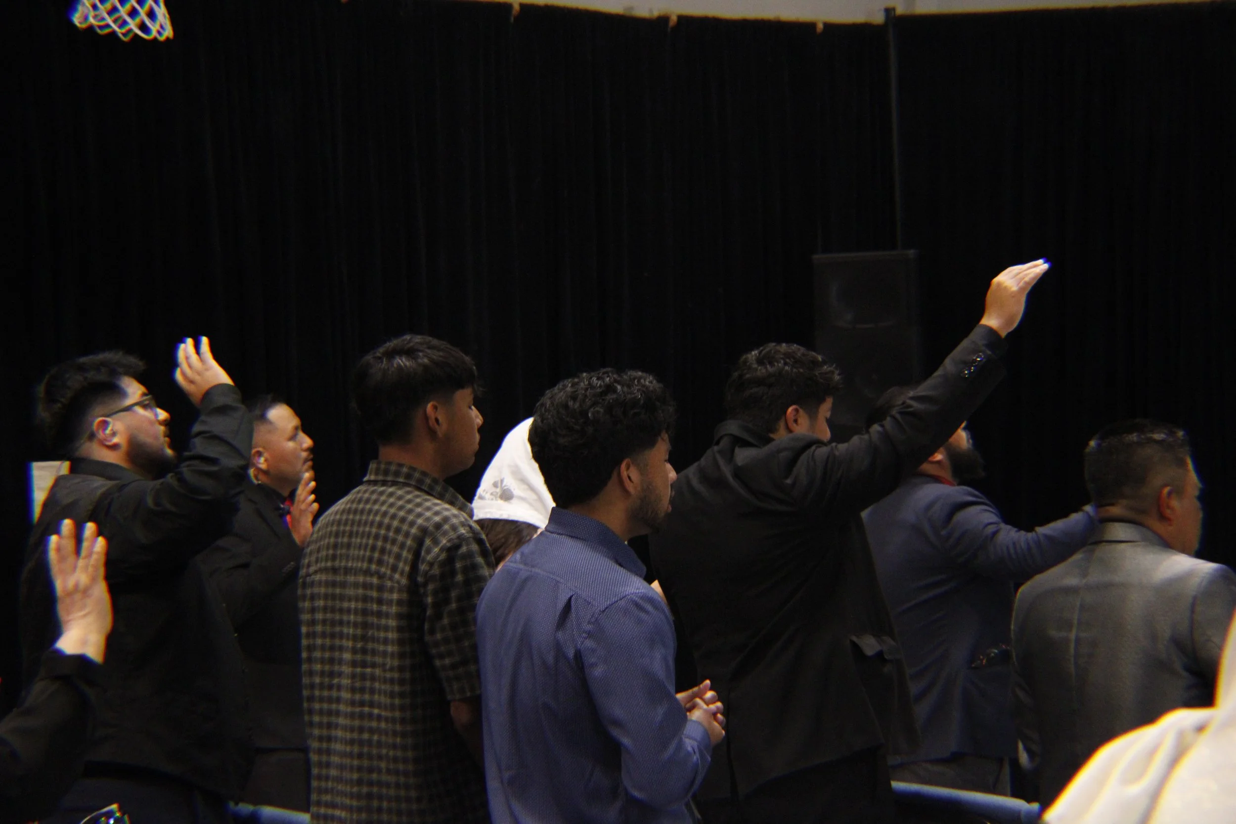A group of people sitting in an auditorium, with some raising their hands, against a dark background with black curtains.