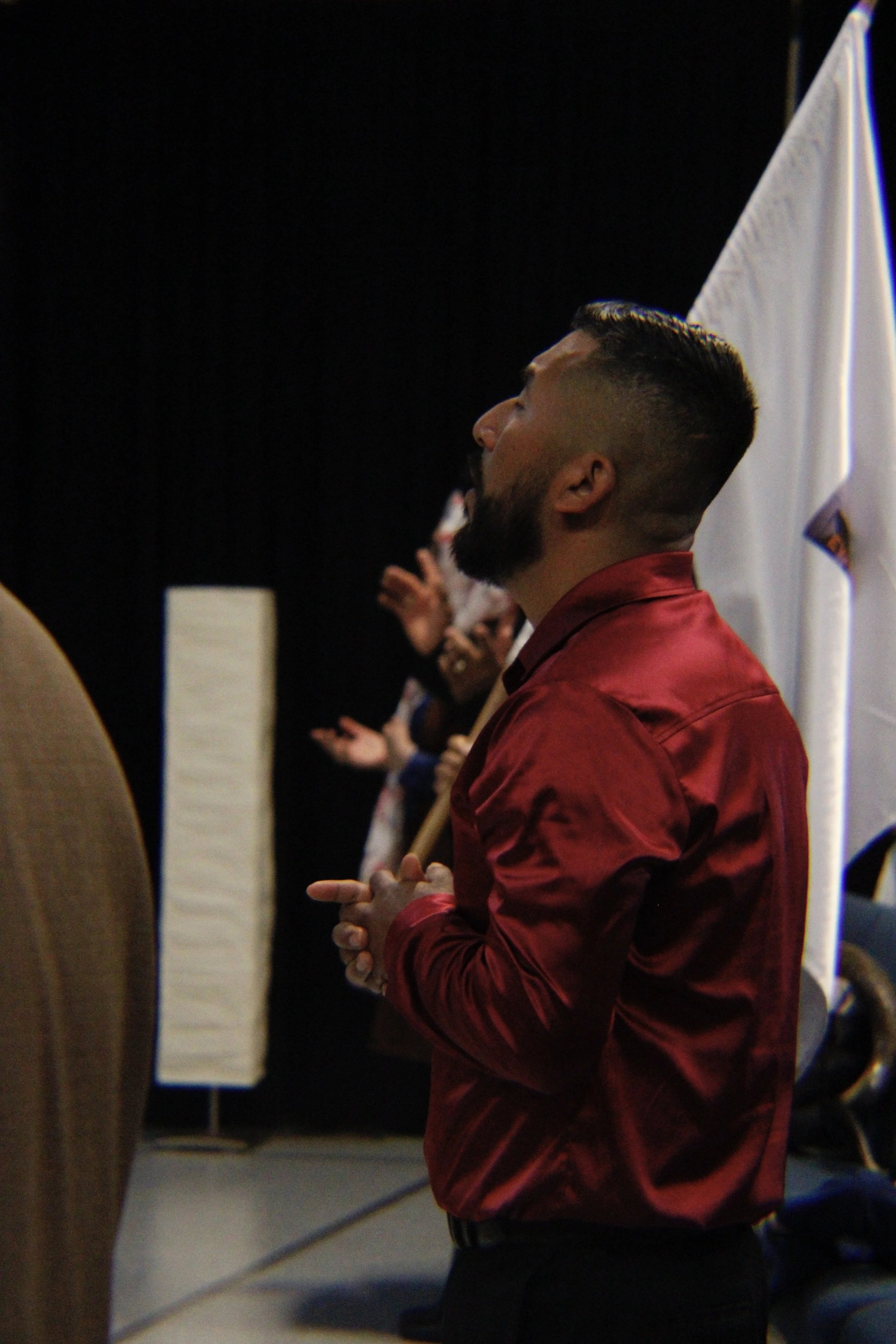 A young man with a beard and a modern haircut, wearing a red silk shirt, holding his hands together, appears to be in a prayer or meditation pose. He is standing inside a building with a dark background, with flags and other people visible behind him.