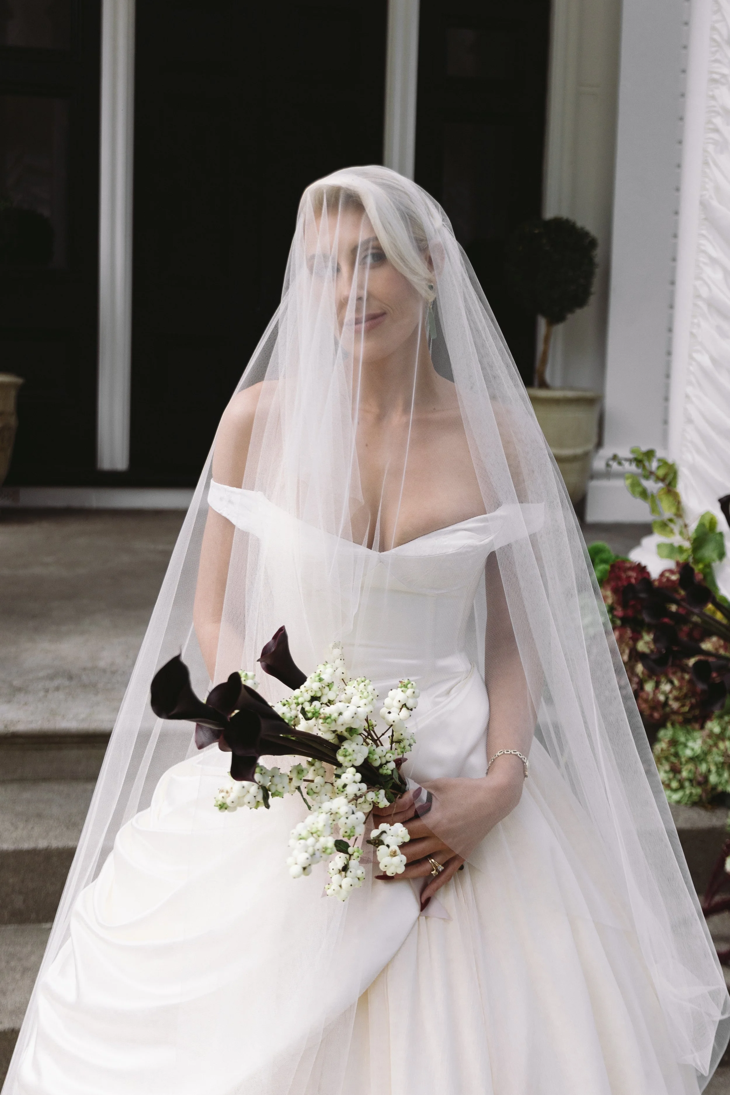 A woman in a white wedding dress with a veil covering her face, holding a bouquet of white and dark purple flowers, standing outside in front of a building with potted plants.