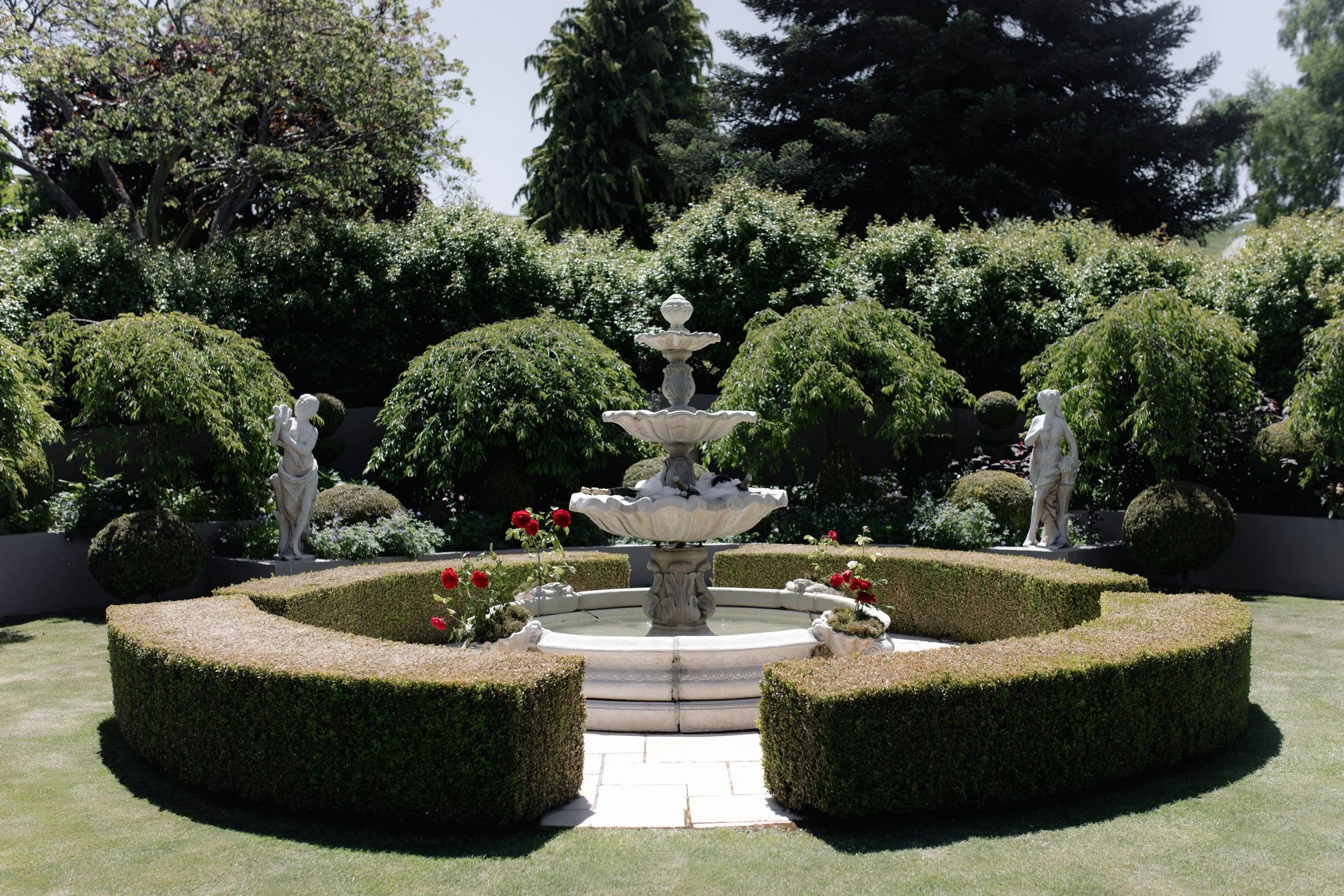 A well-manicured garden with a white marble fountain in the center, surrounded by neatly trimmed bushes and decorative statues of children. There are lush green trees and flowering plants in the background under a clear sky.
