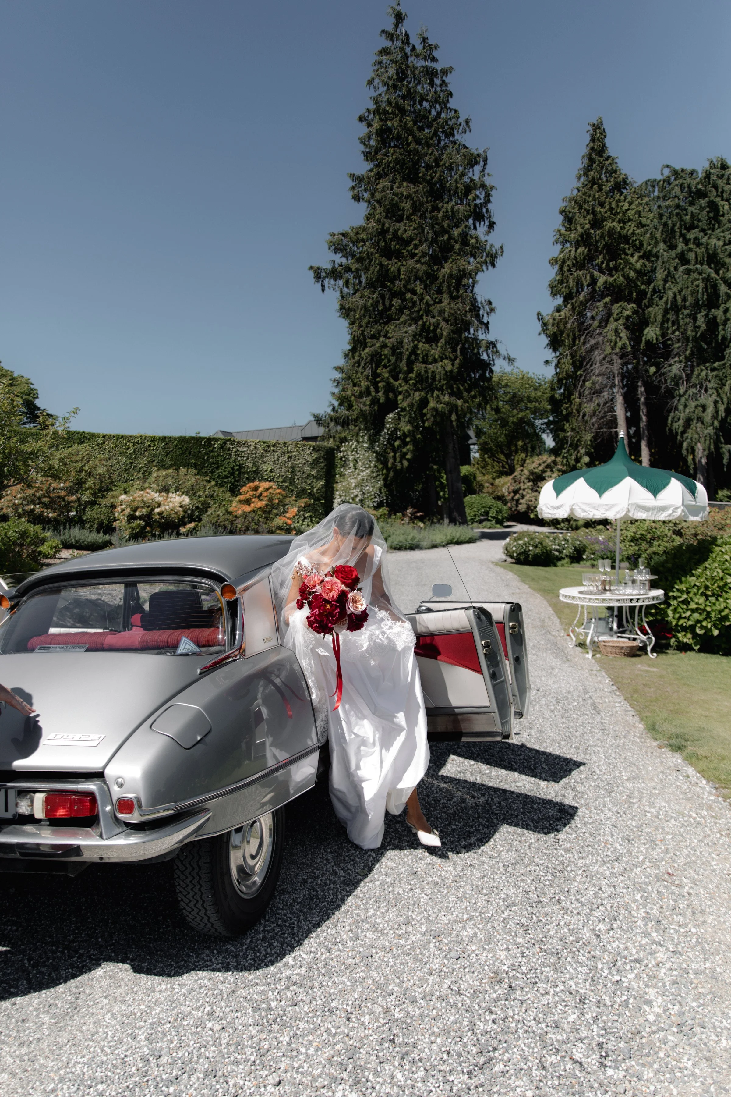 A bride in a wedding dress and veil holding a bouquet of red and pink flowers getting out of a vintage silver car.
