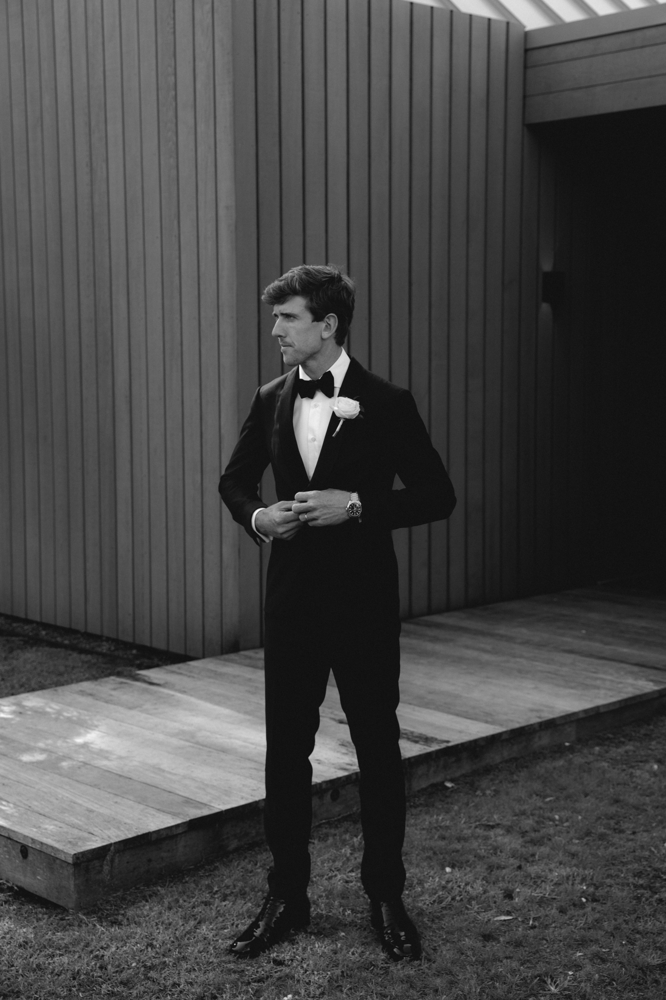 A man in a tuxedo with a bow tie and a flower in his lapel, standing outdoors on a wooden platform next to a modern wooden building, looking to the side with his hands clasped.
