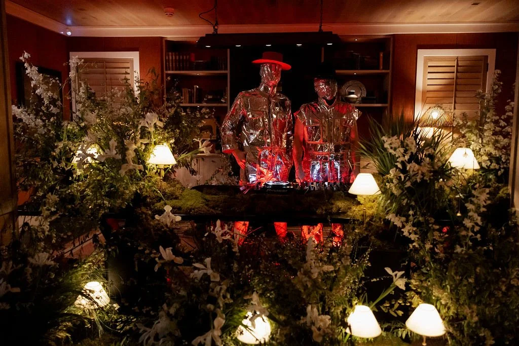 Two metallic mannequins dressed in shiny, reflective clothing with hats, standing among greenery and surrounded by small lamps in a cozy, wooden room.