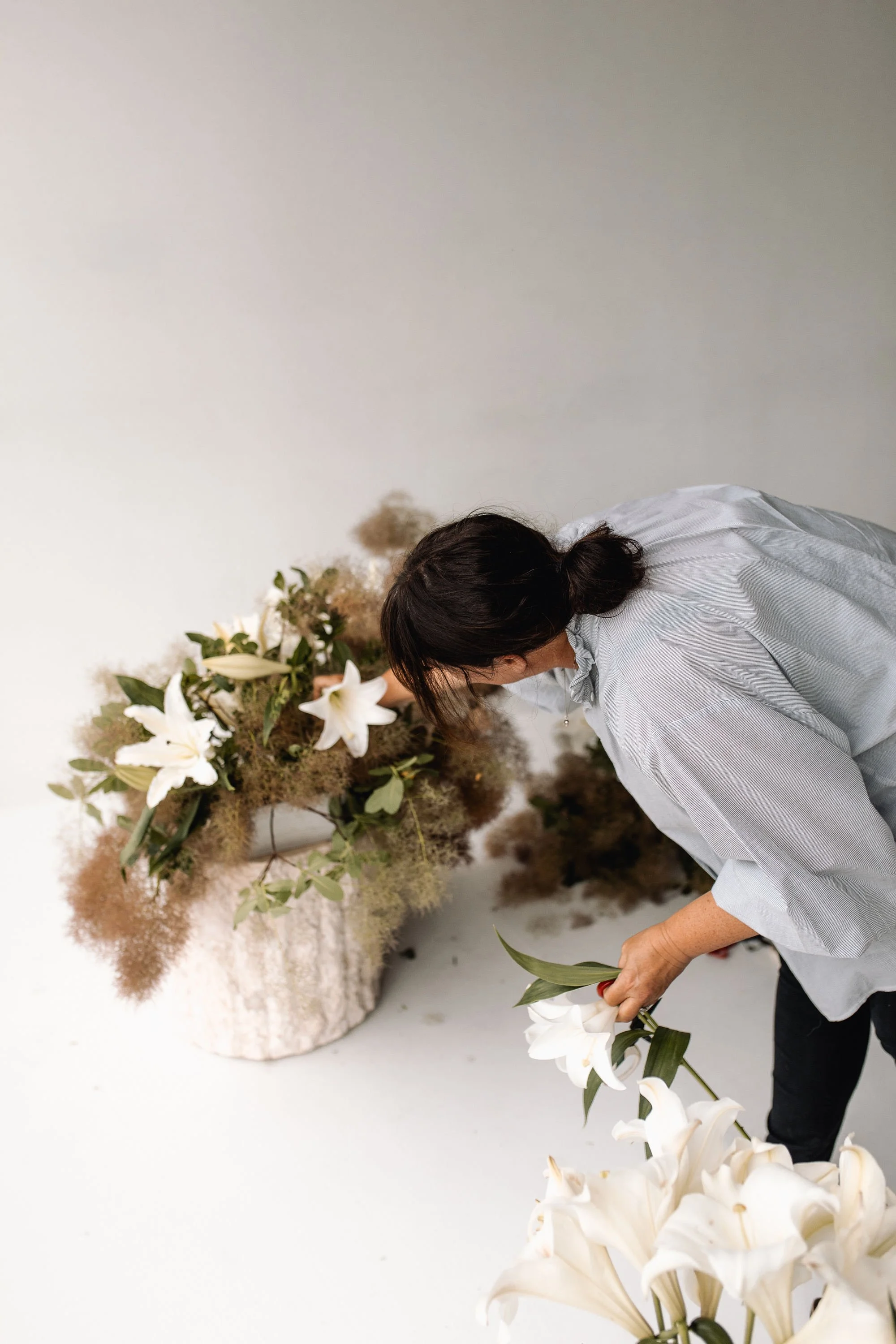 A woman with dark hair wearing a gray striped shirt arranging white lilies in front of a large floral arrangement with white lilies, greenery, and dried pampas grass on a white background.