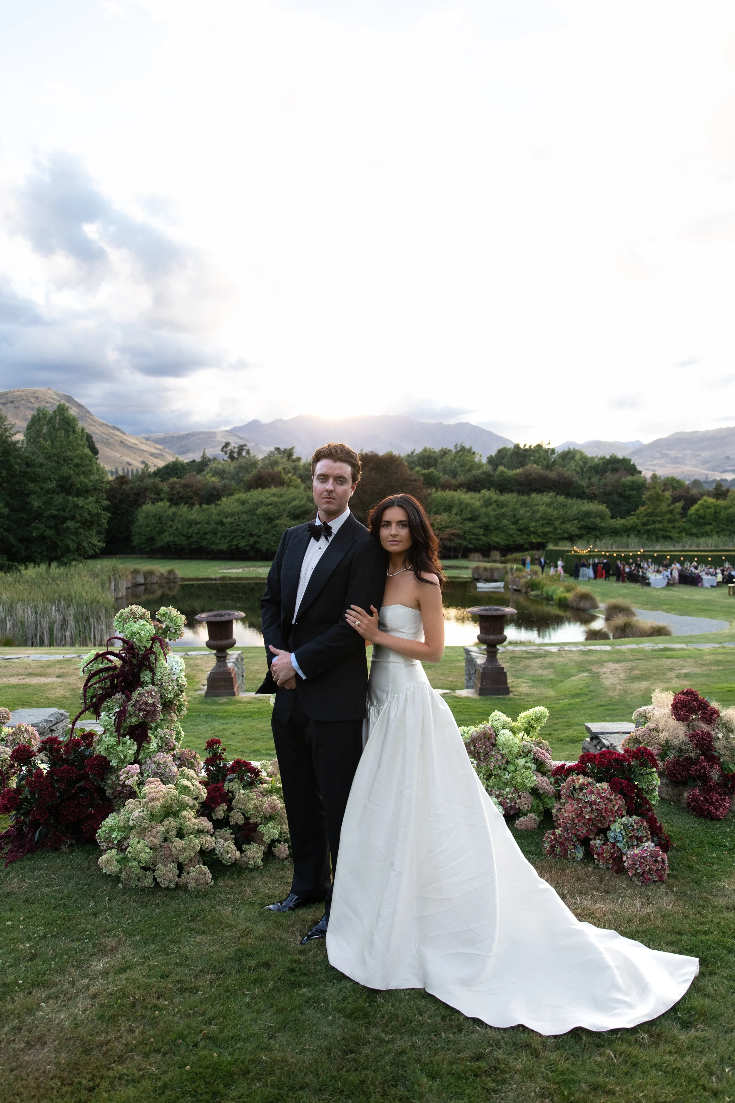 A bride and groom standing together outdoors during a wedding ceremony, with a scenic mountain landscape and a cloudy sky in the background, surrounded by floral arrangements and greenery.