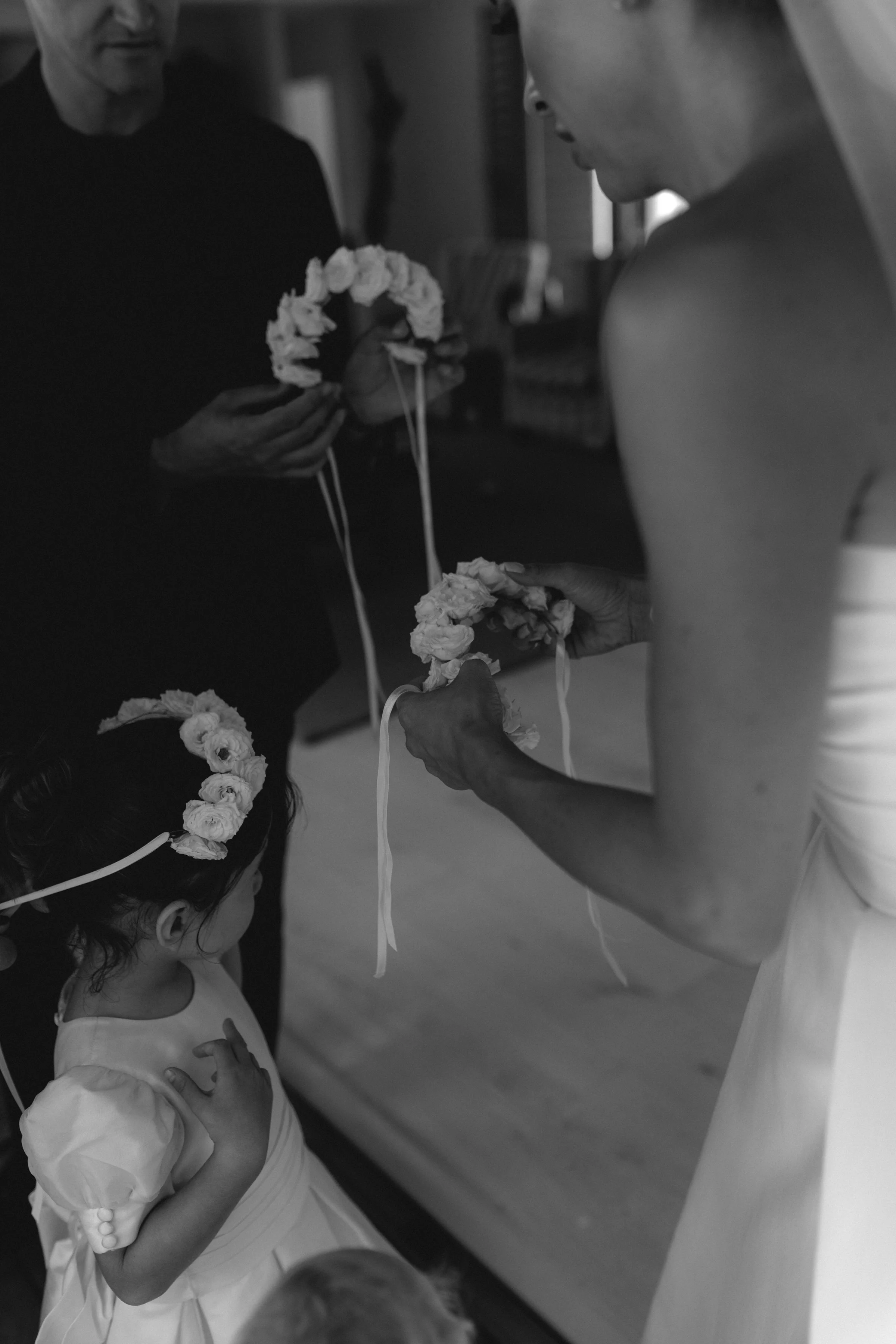 A girl with a flower crown on her head standing with her hand over her heart while looking at a man holding a flower crown, in a room with a blurred background.