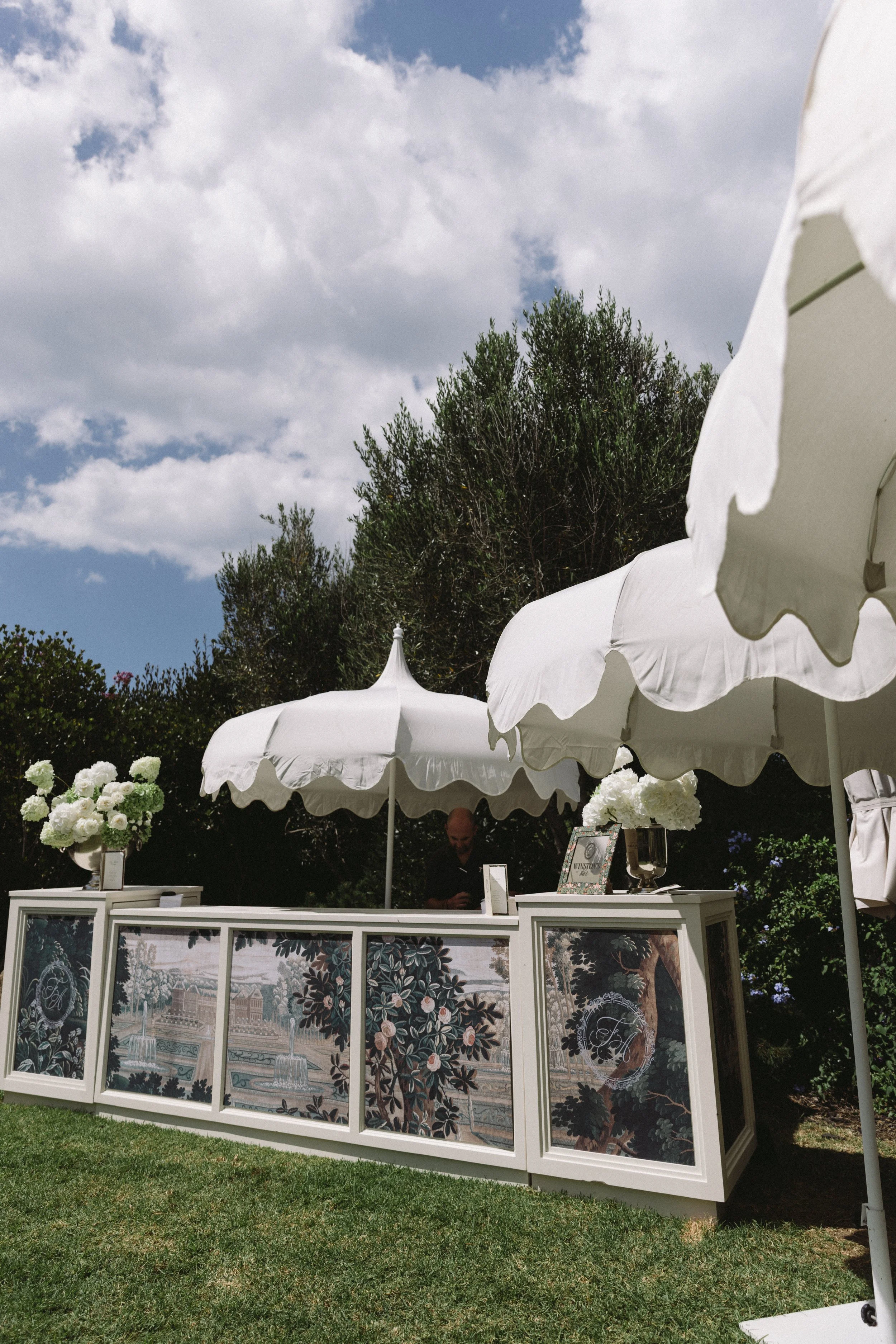 A white flower stand with floral paintings and large white umbrellas on a grassy area, trees, and a cloudy sky in the background.