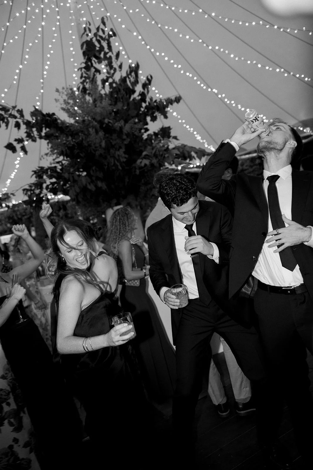 People dancing and drinking at a celebration under a tent decorated with string lights.