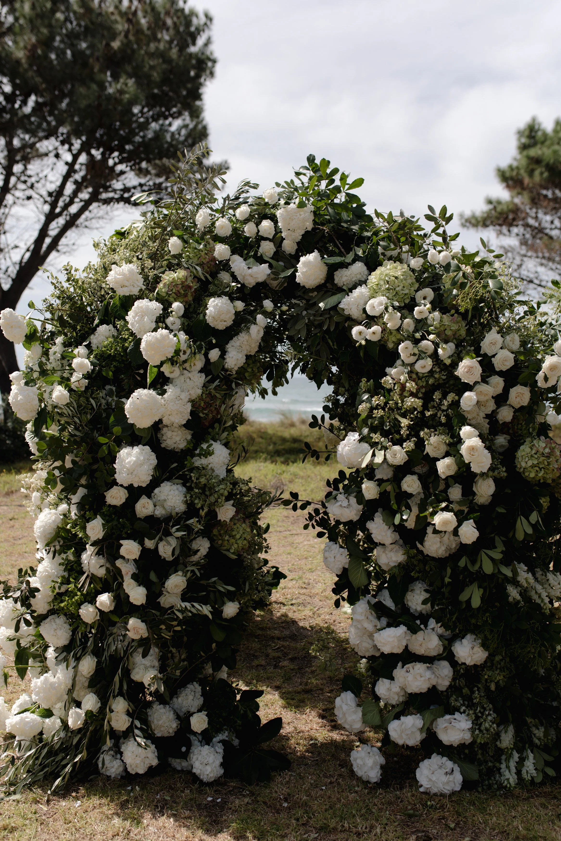 A floral arch made of white roses, hydrangeas, and greenery, set outdoors on a grassy area with trees and a cloudy sky in the background, typically used for wedding ceremonies.