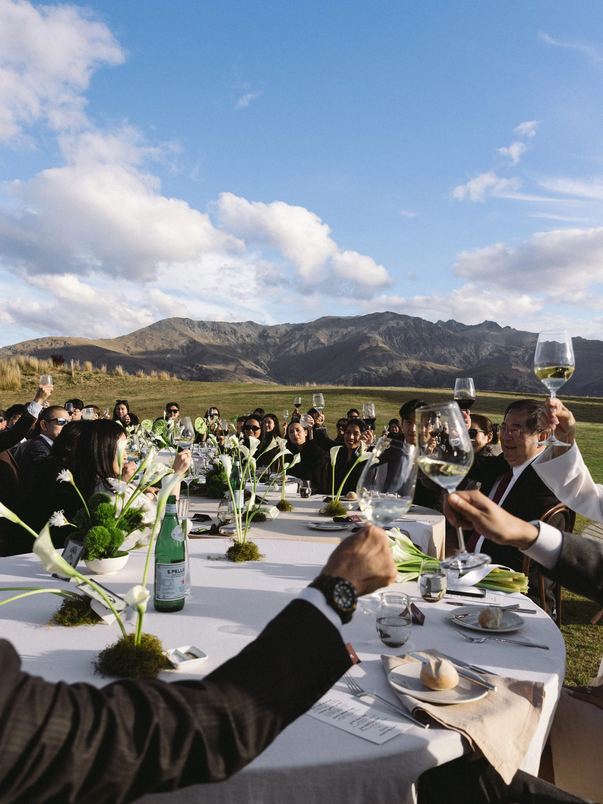 People at a large outdoor dining event holding up glasses of white wine in a toast, with mountainous landscape and blue sky in the background.