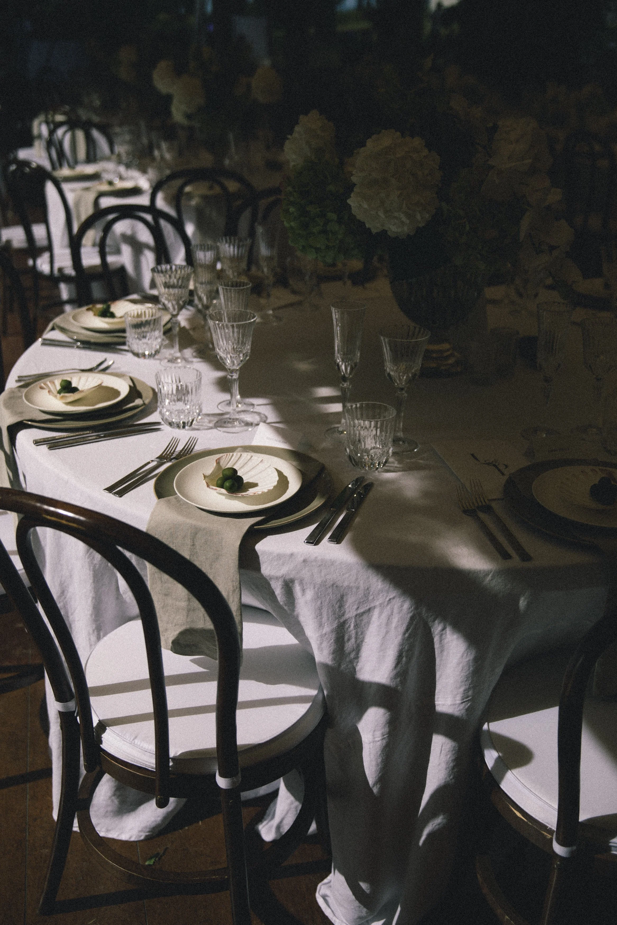 A long dining table covered with a white tablecloth, set with white plates, silverware, and clear glassware, arranged for a formal meal. Empty black chairs surround the table, and a large floral centerpiece with white and green flowers is visible at the center. The setting appears to be in a dimly lit room or area.