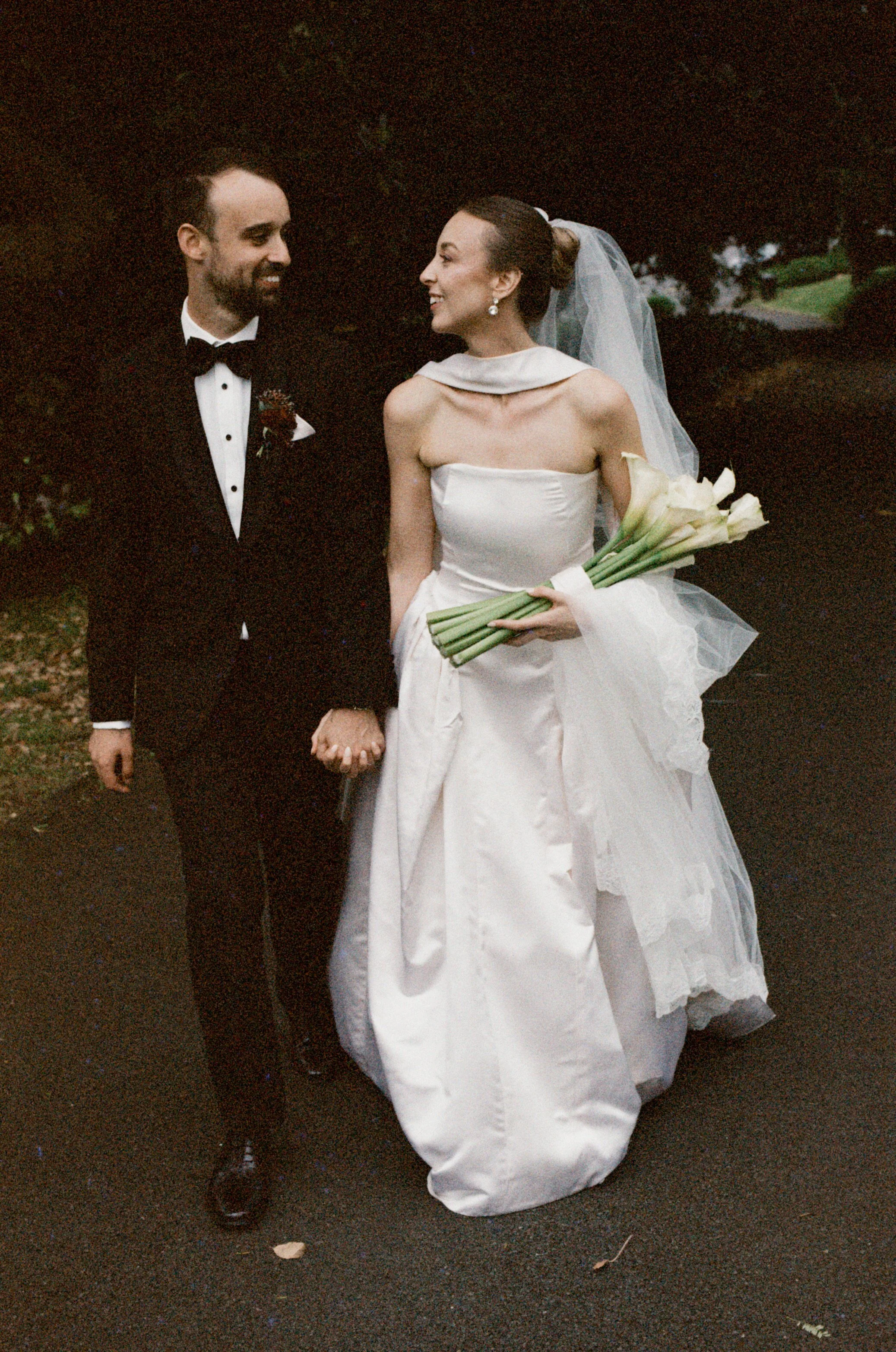 A bride and groom holding hands outdoors during a wedding, with the bride in a white gown and holding a bouquet of calla lilies, while the groom wears a black tuxedo with a bow tie.
