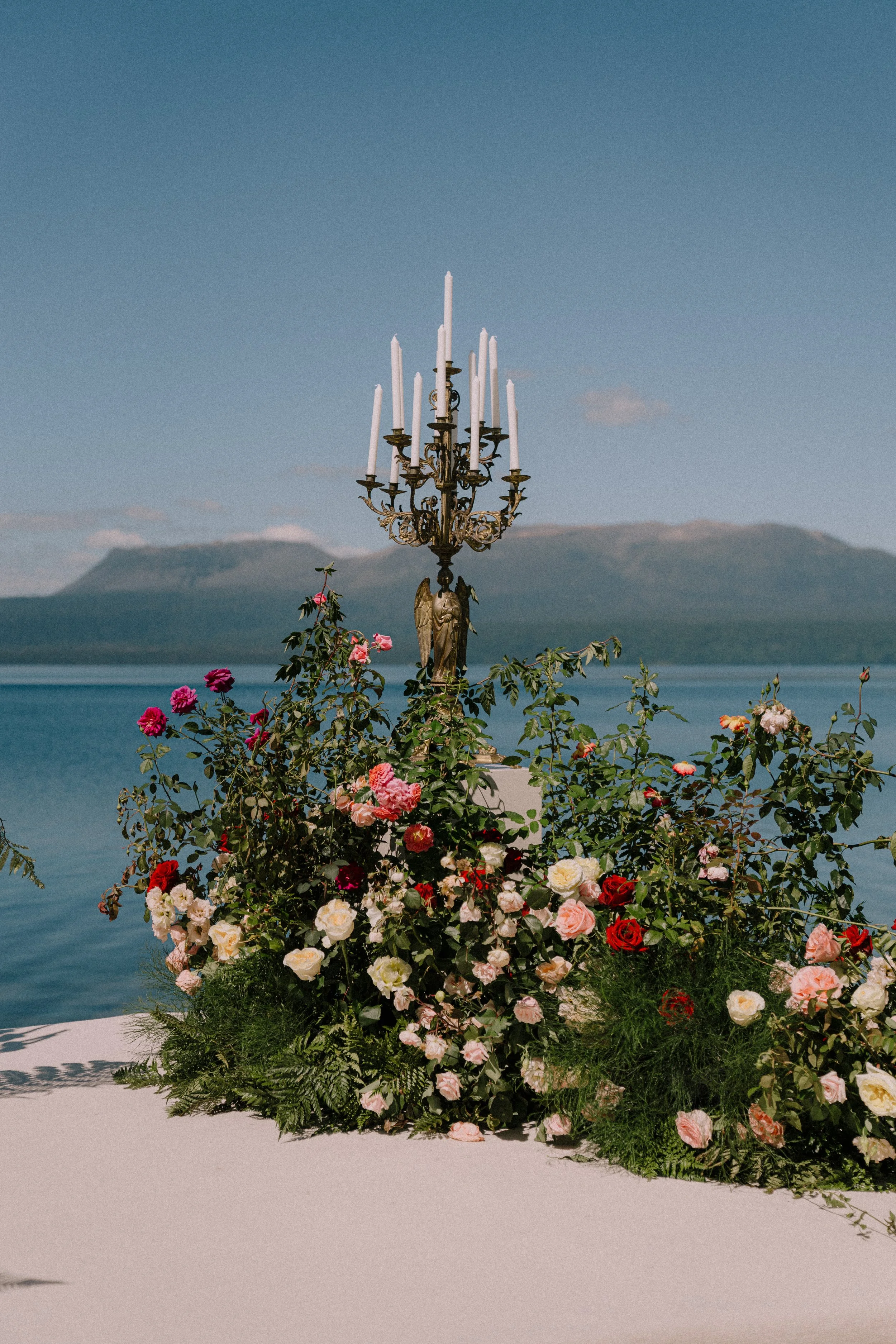 A decorative street lamp with multiple candles, surrounded by colorful roses and greenery, with a lake and mountains in the background.