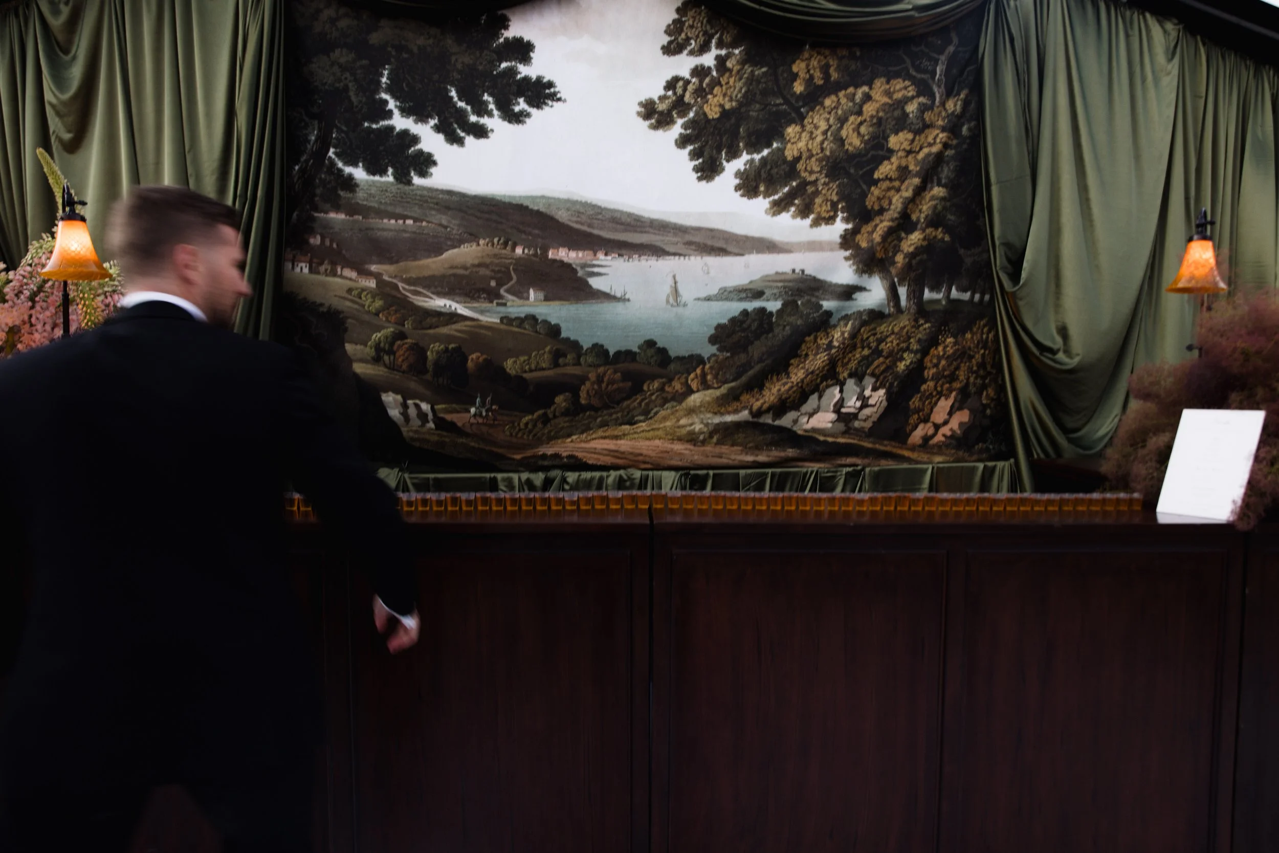 Painting of a landscape view with water, trees, hills, boats, and a cloudy sky, surrounded by green curtains and lit by two small table lamps.