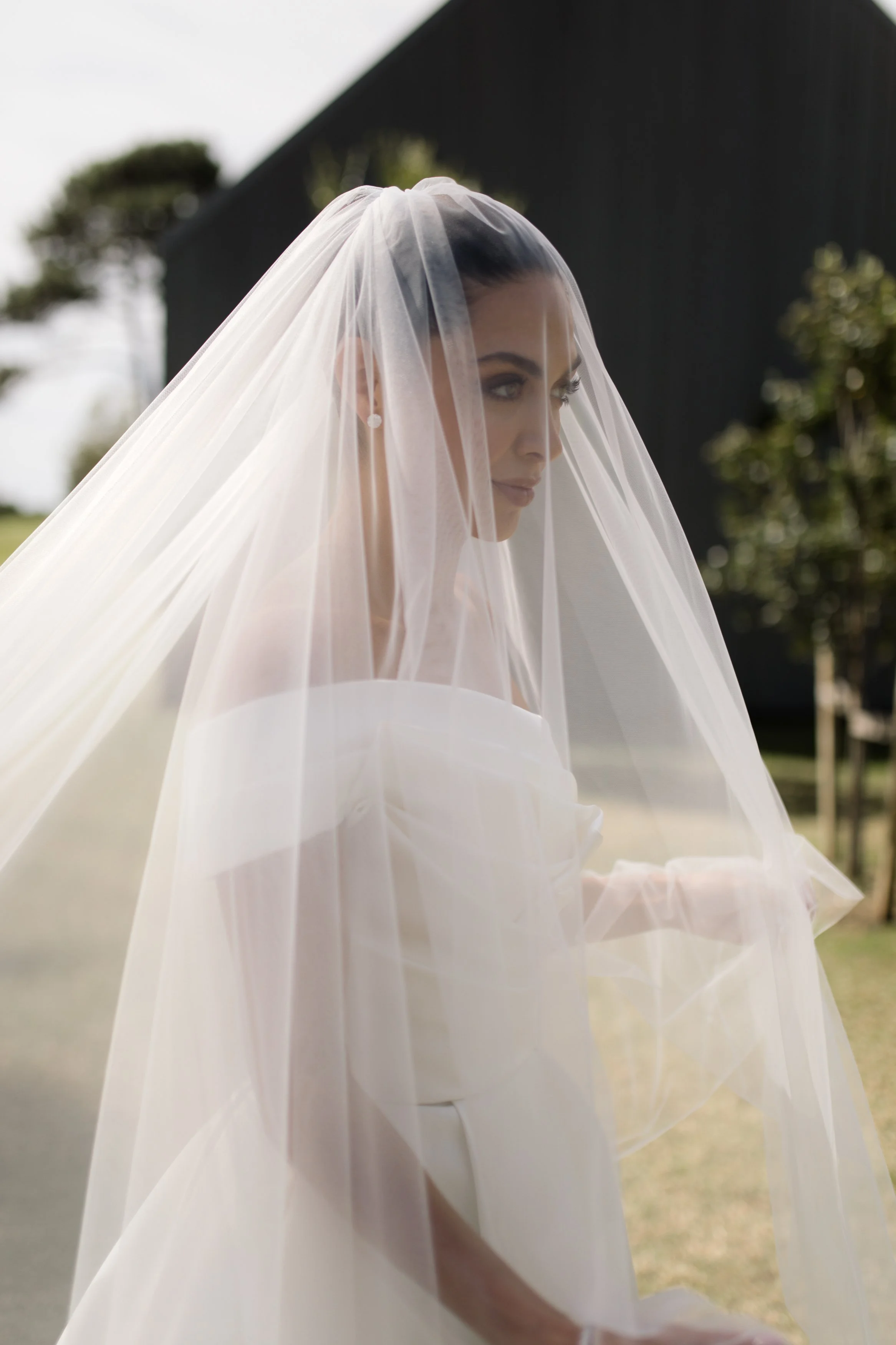 A bride with dark hair in a bun, wearing a white wedding dress and veil, outdoors with trees and a dark building in the background.