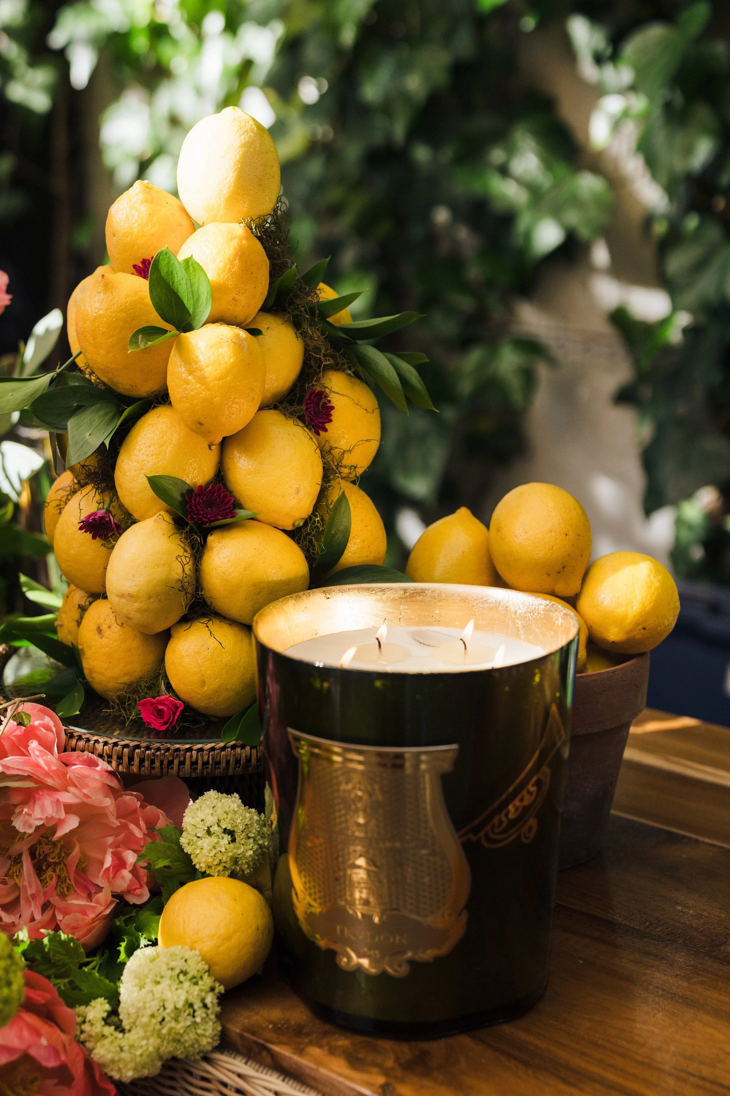 A floral arrangement of yellow lemons with green leaves and small red and pink flowers, alongside a lit candle in a metallic holder, on a wooden table surrounded by additional flowers and greenery.